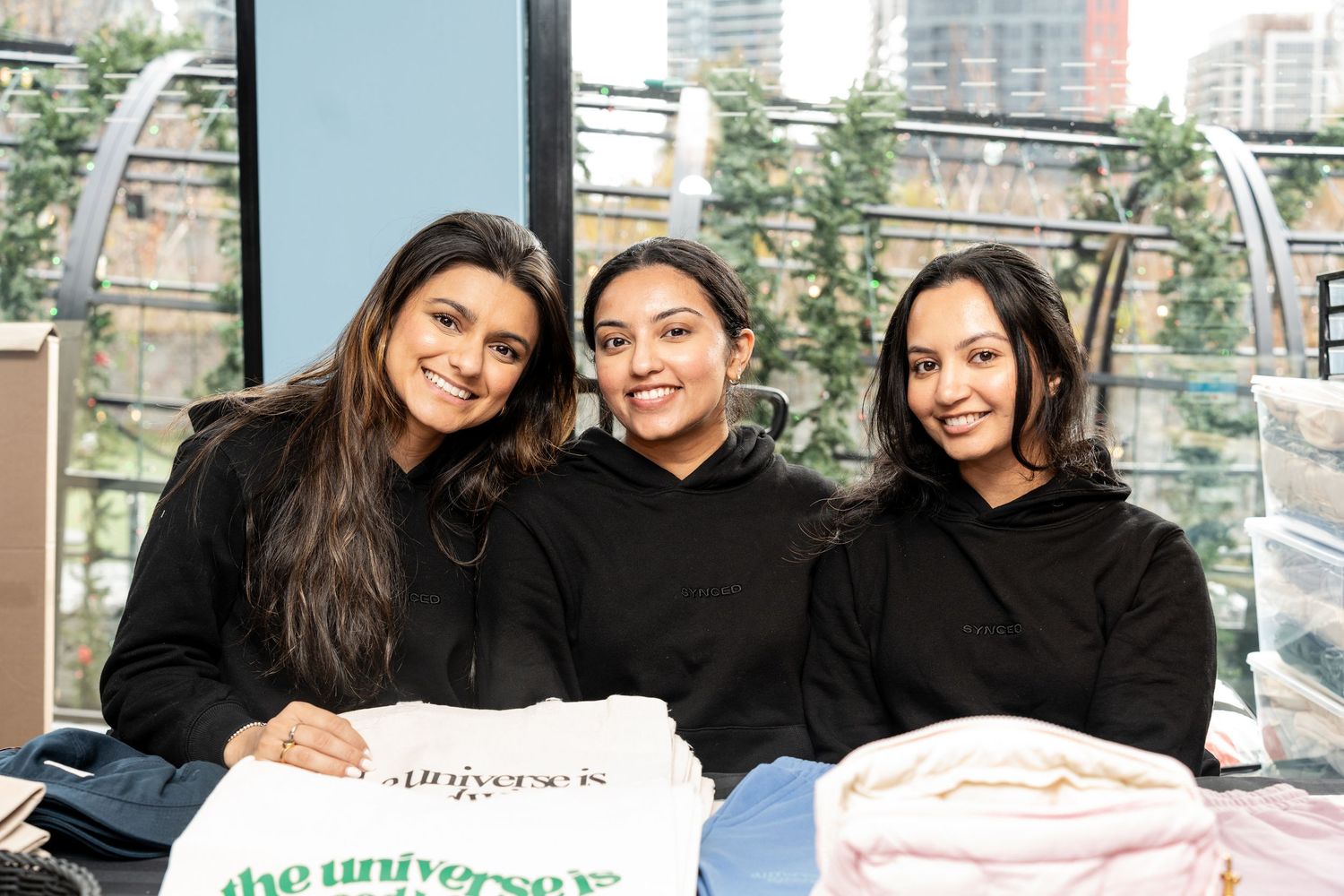 Three smiling young women posing together at a vendor booth with folded merchandise at a local market.