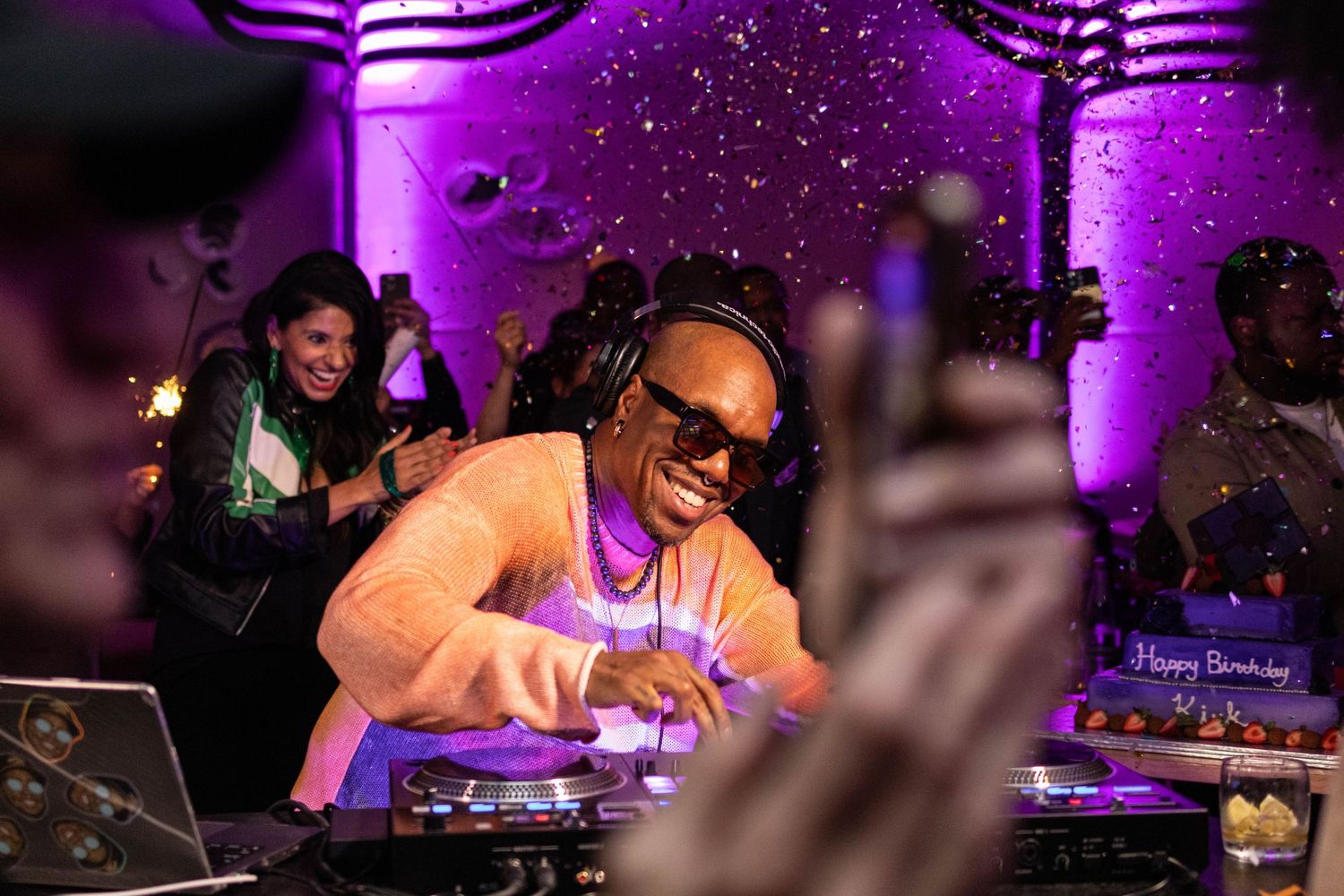 DJ wearing sunglasses and headphones performing at a lively birthday party with purple lighting.