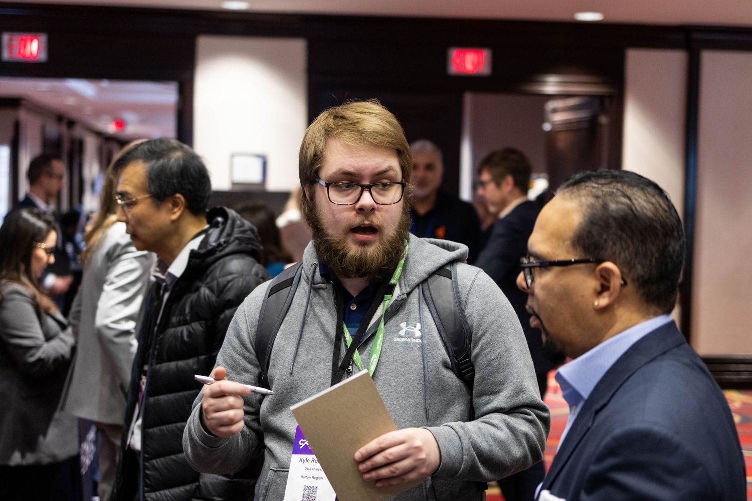 Young bearded man with conference lanyard holding clipboard in conversation at professional networking event.