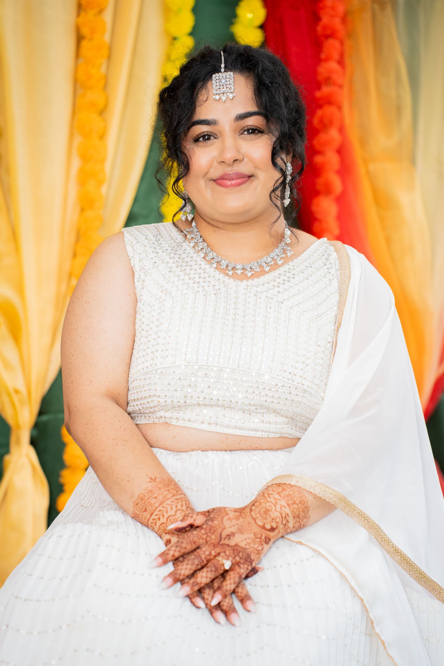 Indian bride in white lehenga with diamond jewelry and mehndi hands at Haldi ceremony.