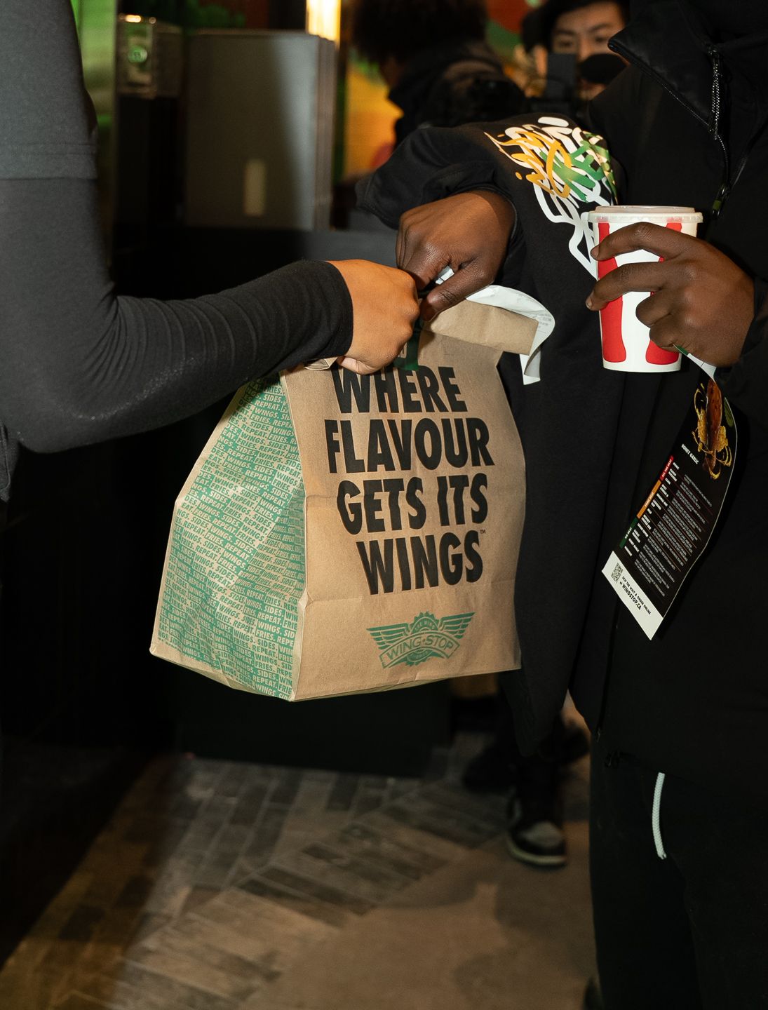 Close-up of person receiving branded paper bag reading Where Flavour Gets Its Wings at restaurant opening.