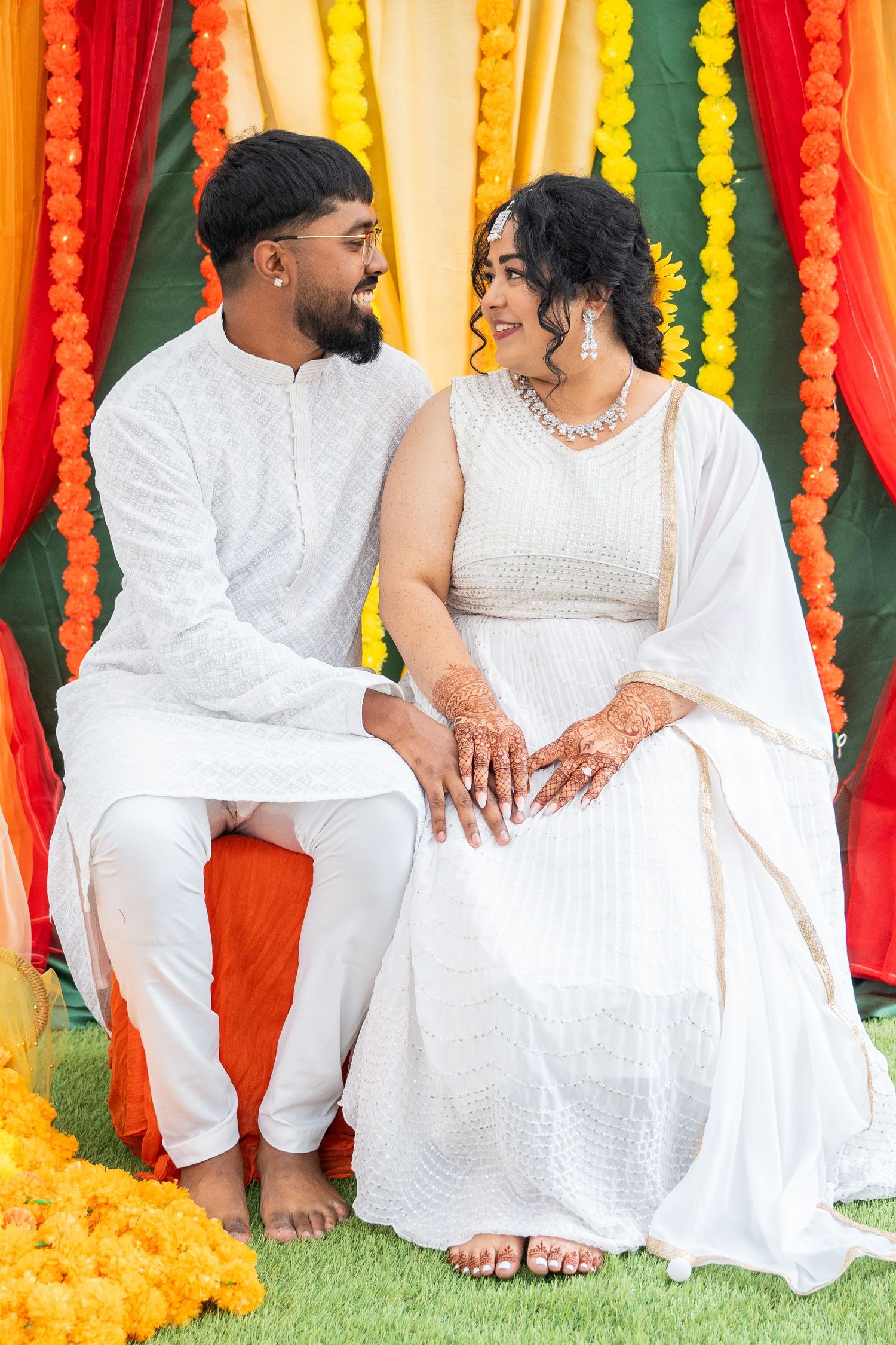 Bride and groom in white outfits sitting together at a colorful Indian Haldi ceremony backdrop.