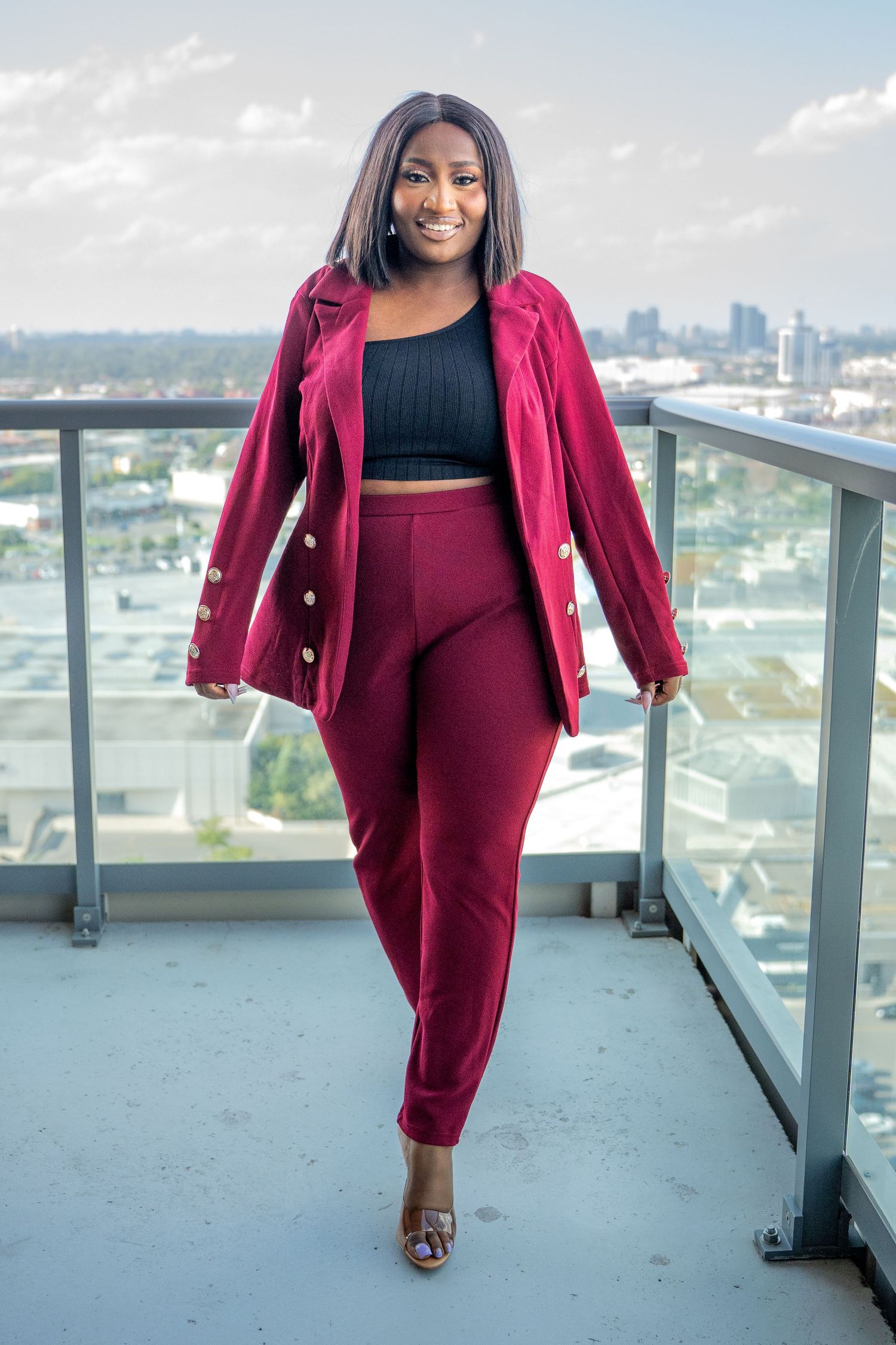 Stylish woman in burgundy blazer and pants posing on a rooftop with city skyline view.