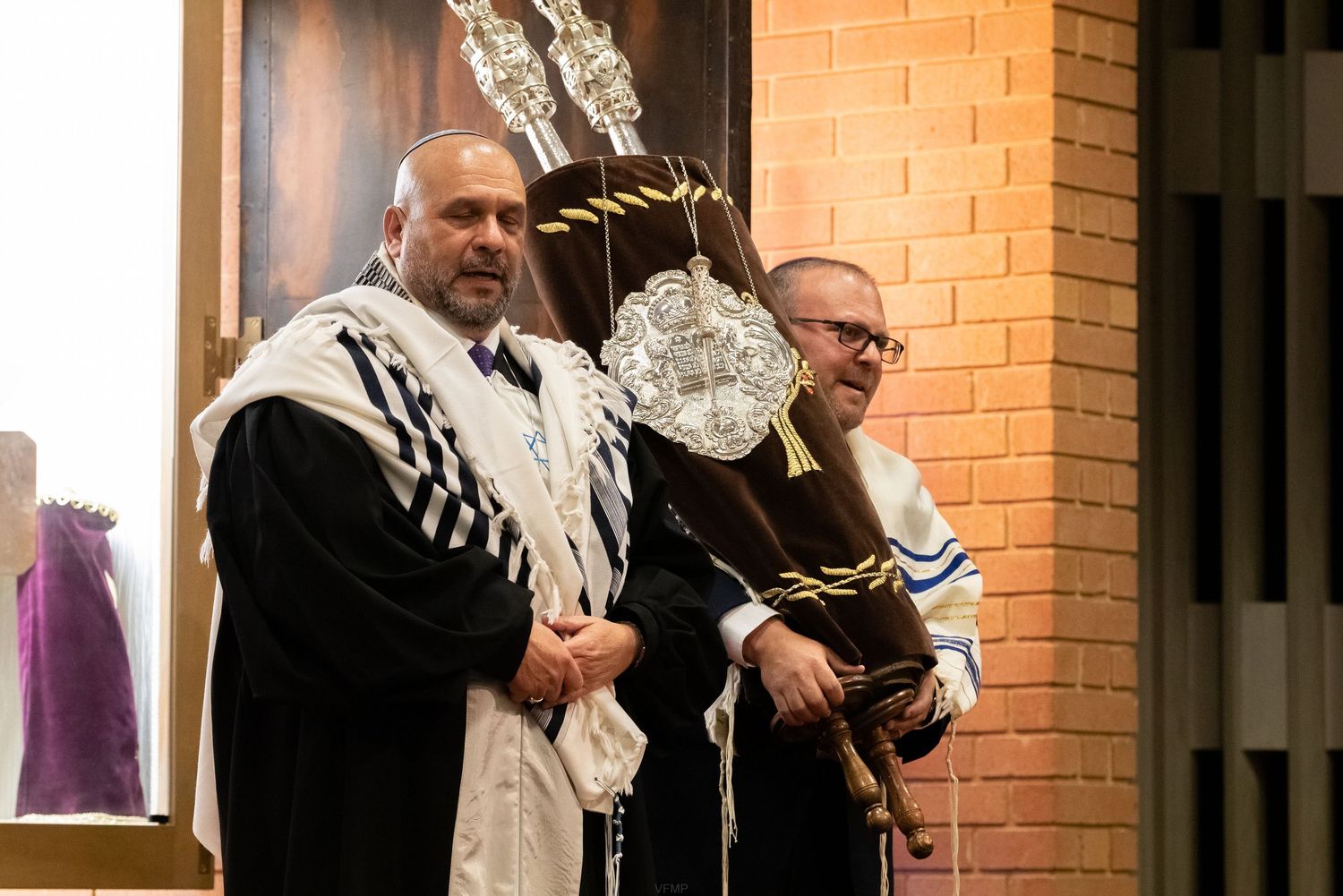 Two people in religious clothing greet each other warmly at the entrance of a brick building.