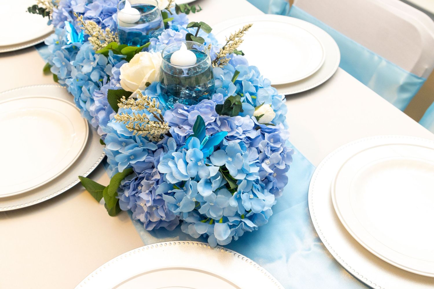 Blue hydrangea centerpiece with white plates and candles on an elegant table setting for a special occasion.