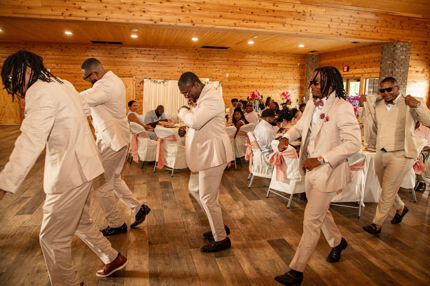 Groomsmen in white suits dancing energetically at wedding reception in rustic venue.