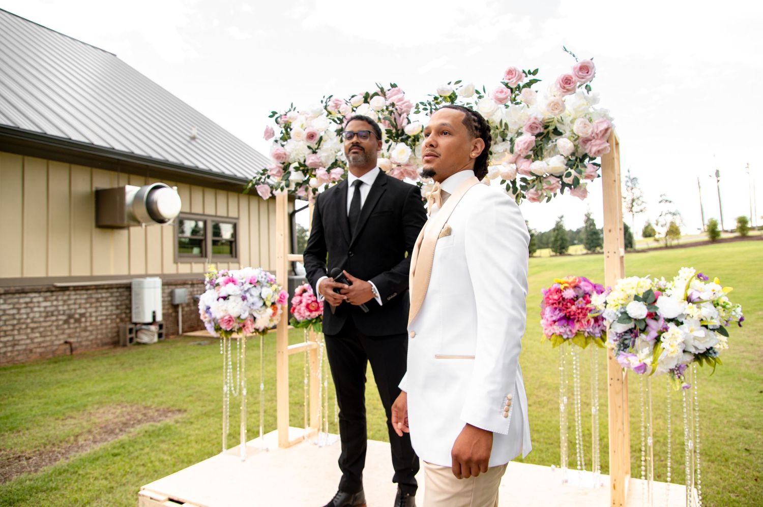 Groom in white suit and groomsman standing at outdoor floral wedding altar.