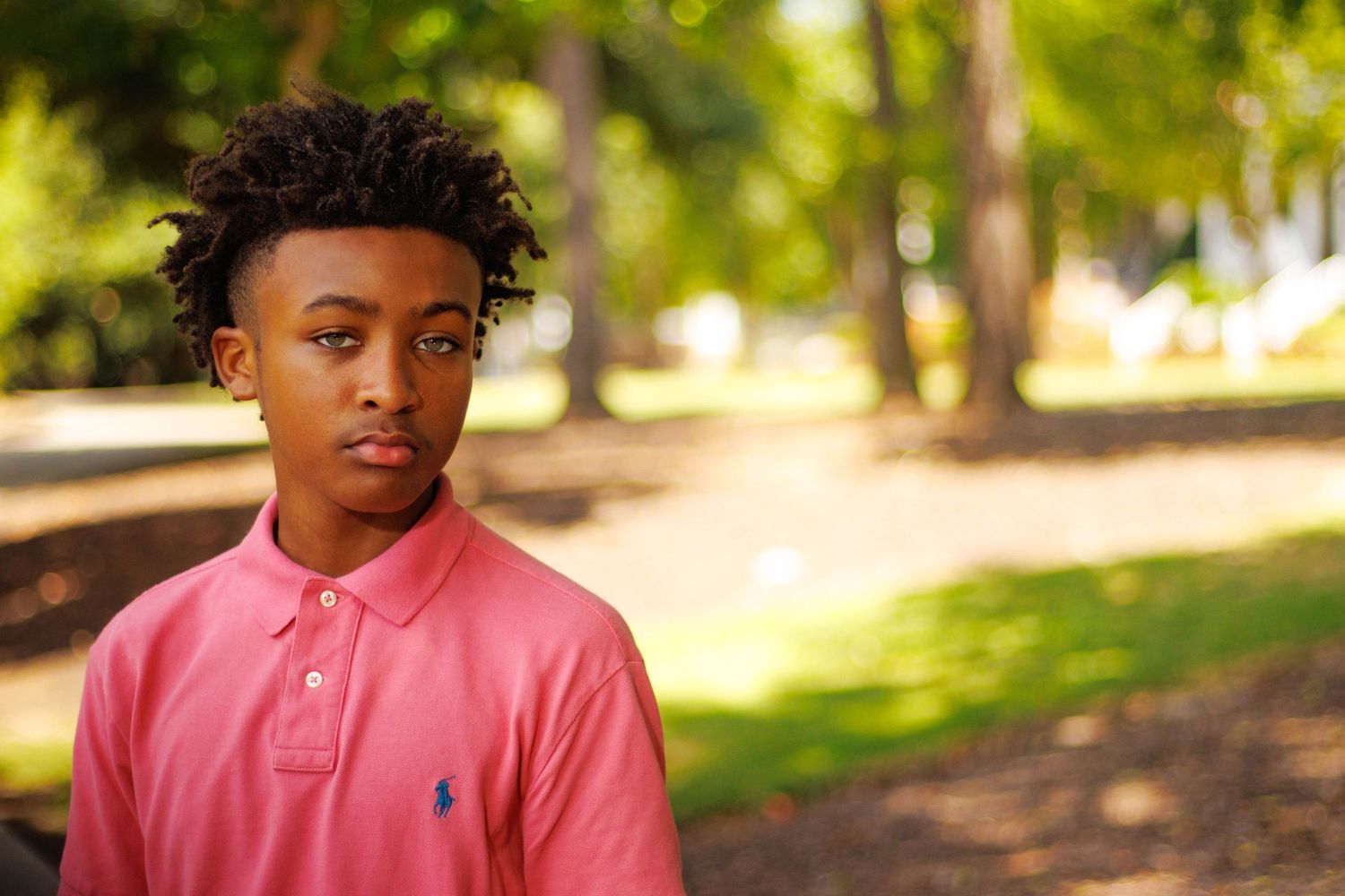 Young man in pink Ralph Lauren polo shirt standing outdoors in a green park setting.