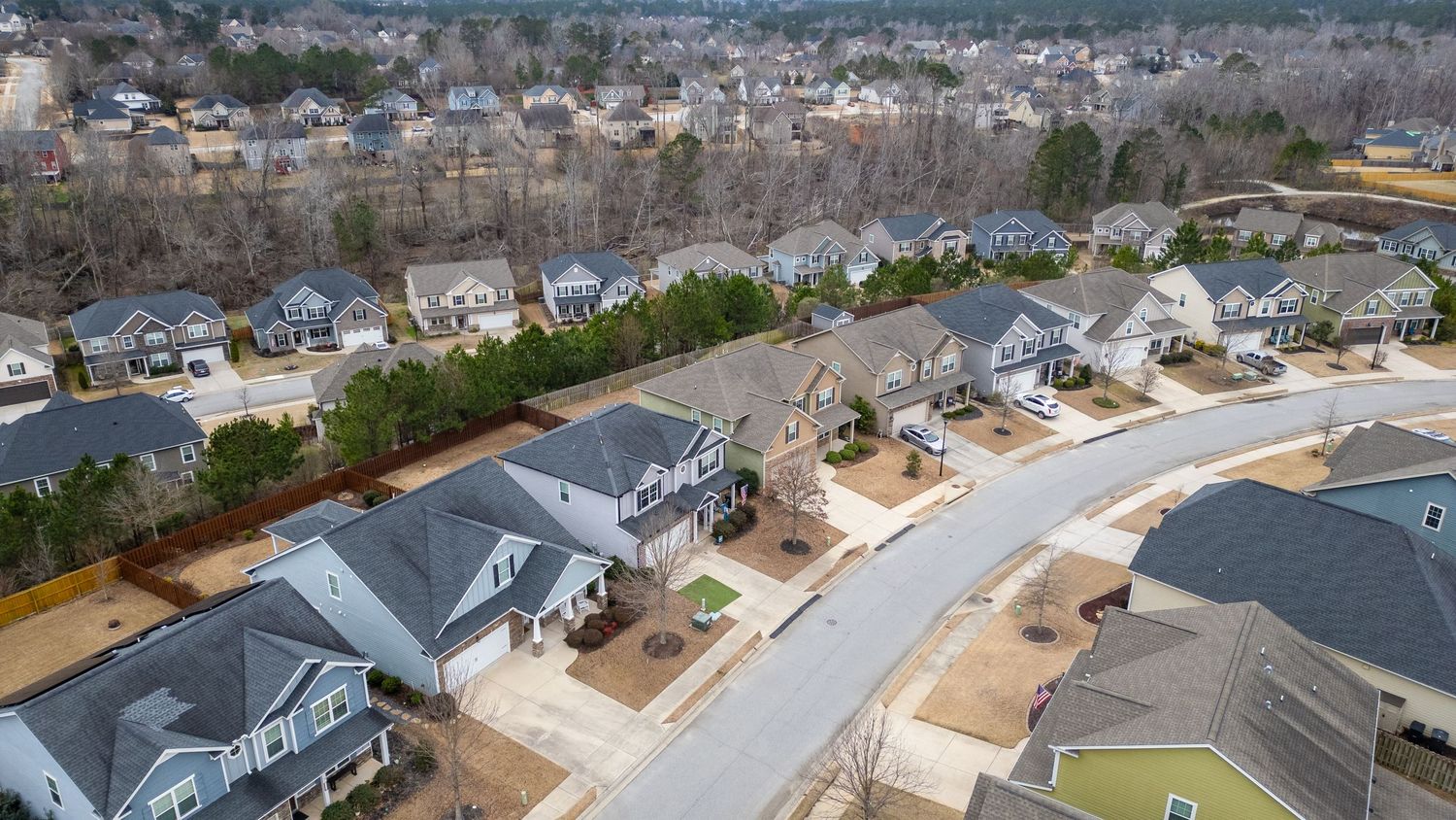 Aerial view of a suburban neighborhood with curved streets, modern homes, and gray rooftops surrounded by bare winter trees.