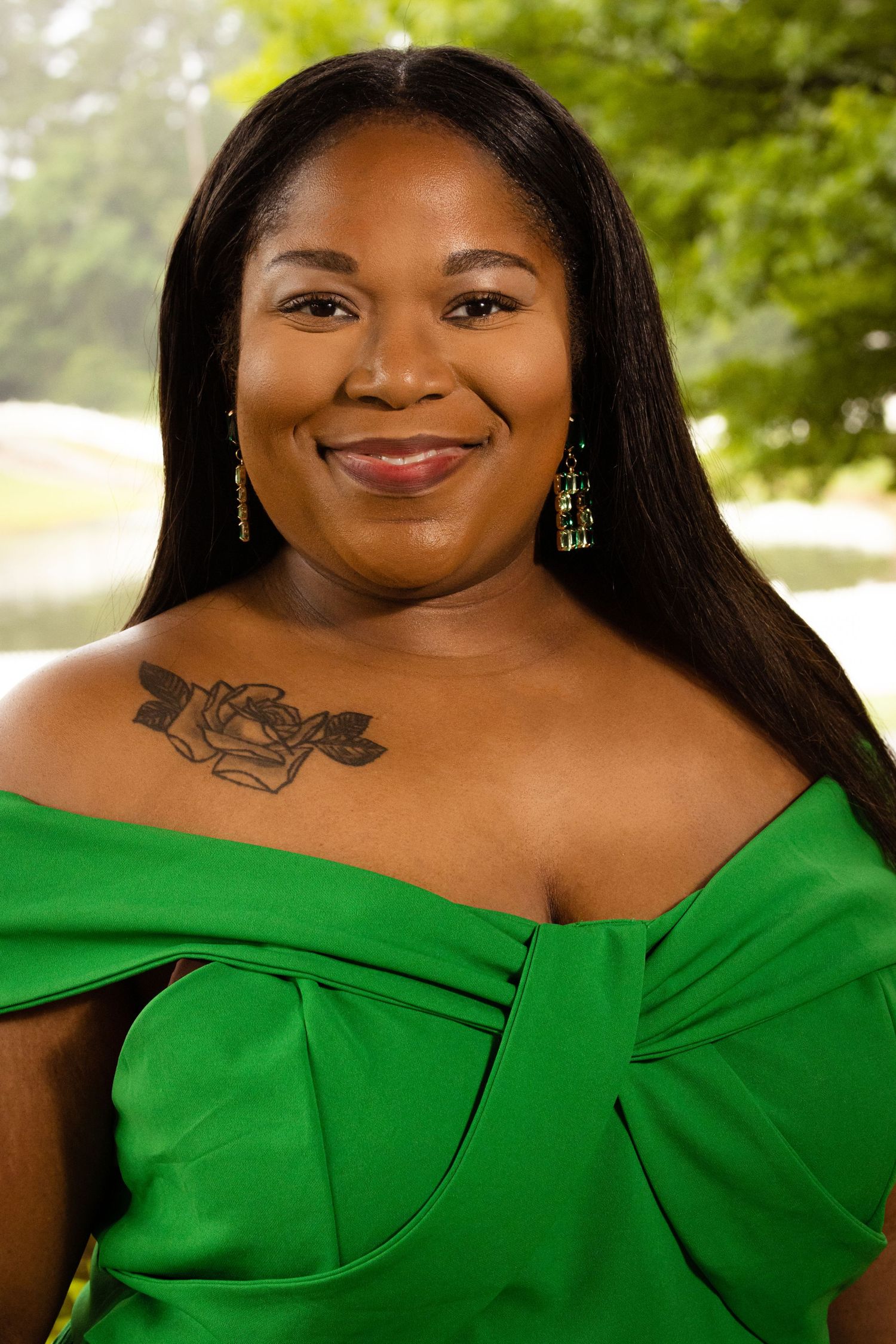 Woman in green off-shoulder dress with rose chest tattoo smiles outdoors near trees.