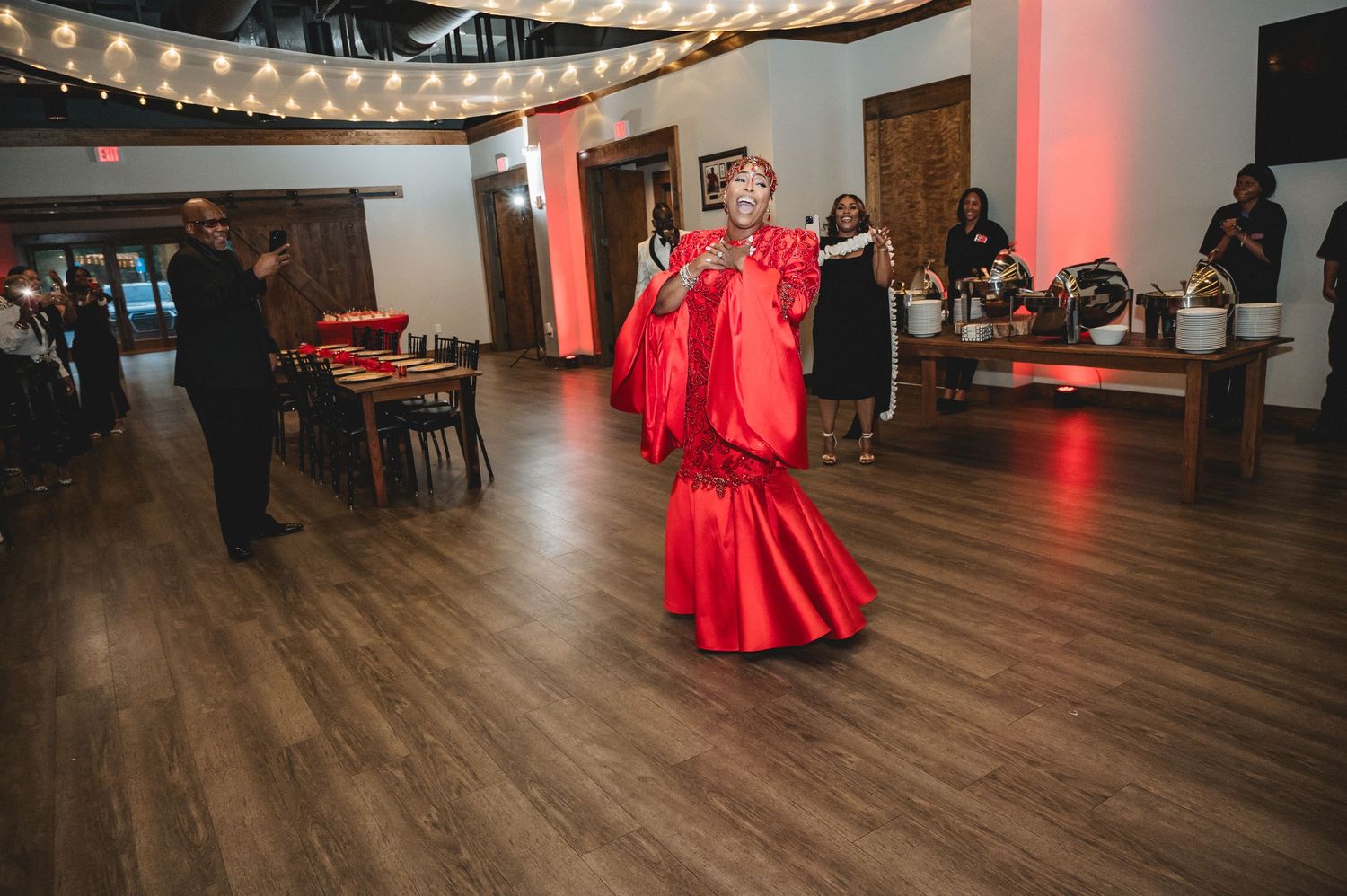String lights illuminate a party venue with red decorative elements and hardwood flooring.