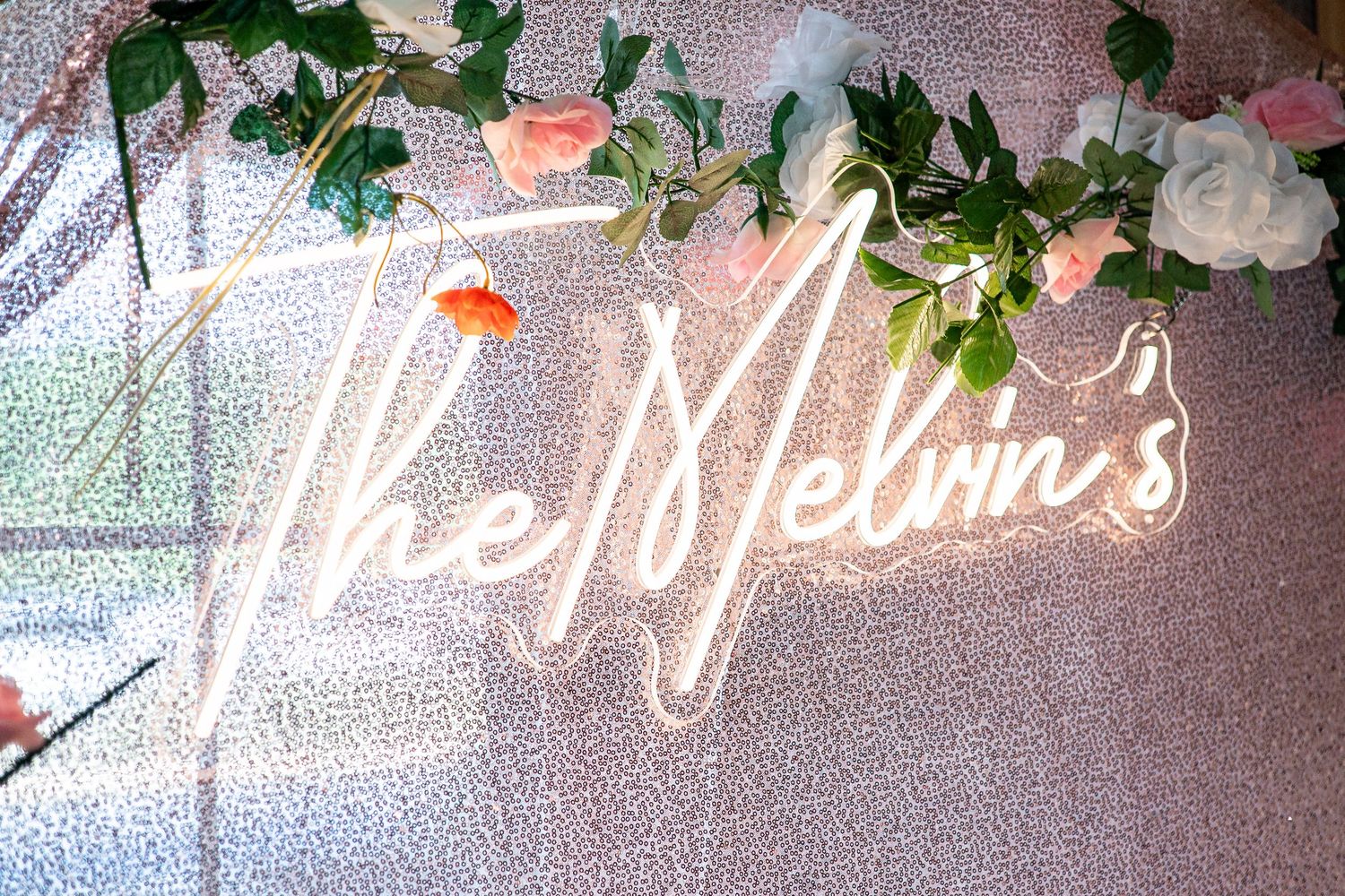 Neon sign reading The Melvins glows pink against sequin backdrop decorated with flowers at wedding.