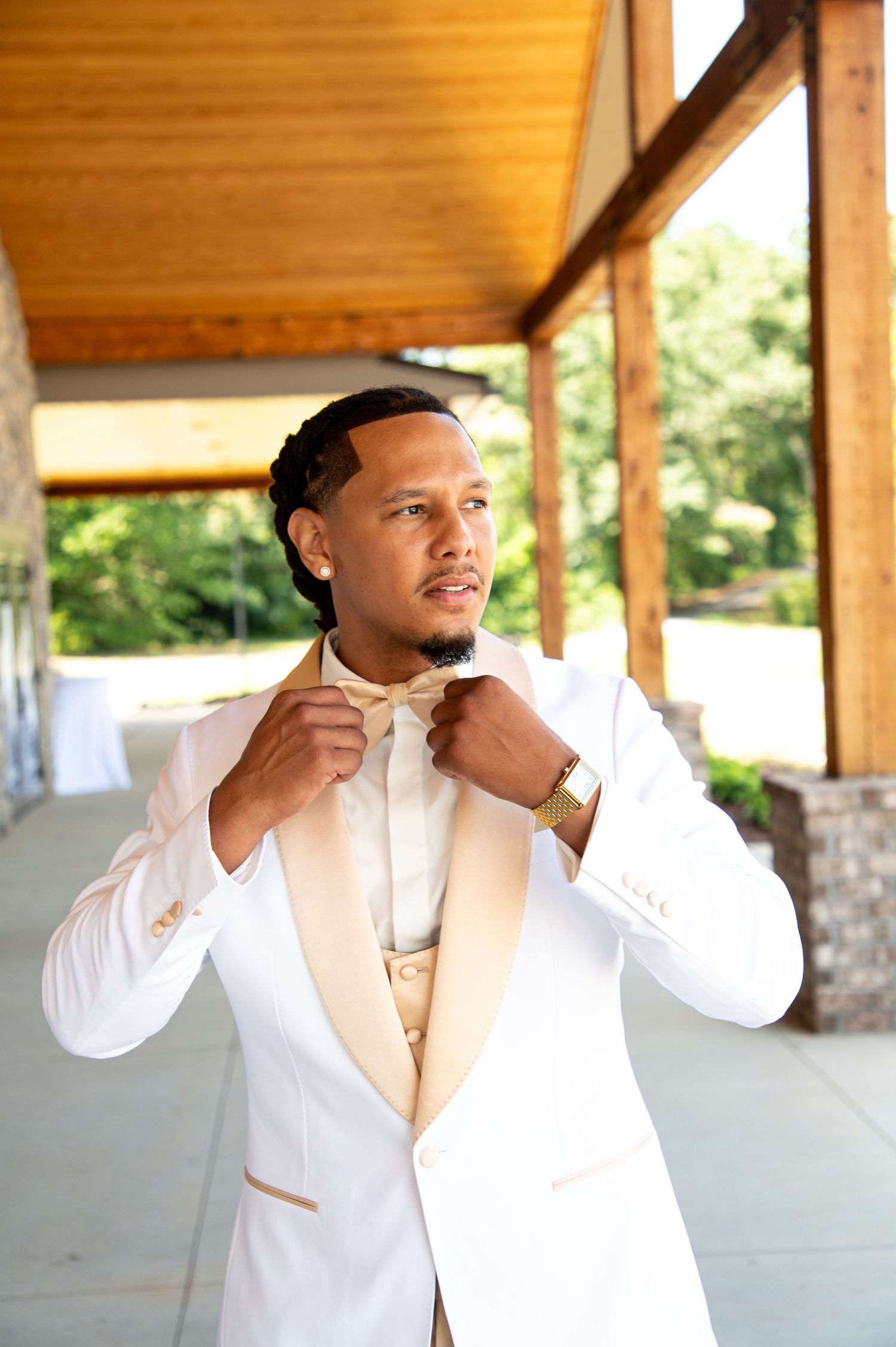 Groom in white tuxedo adjusting bow tie outdoors at wedding venue.