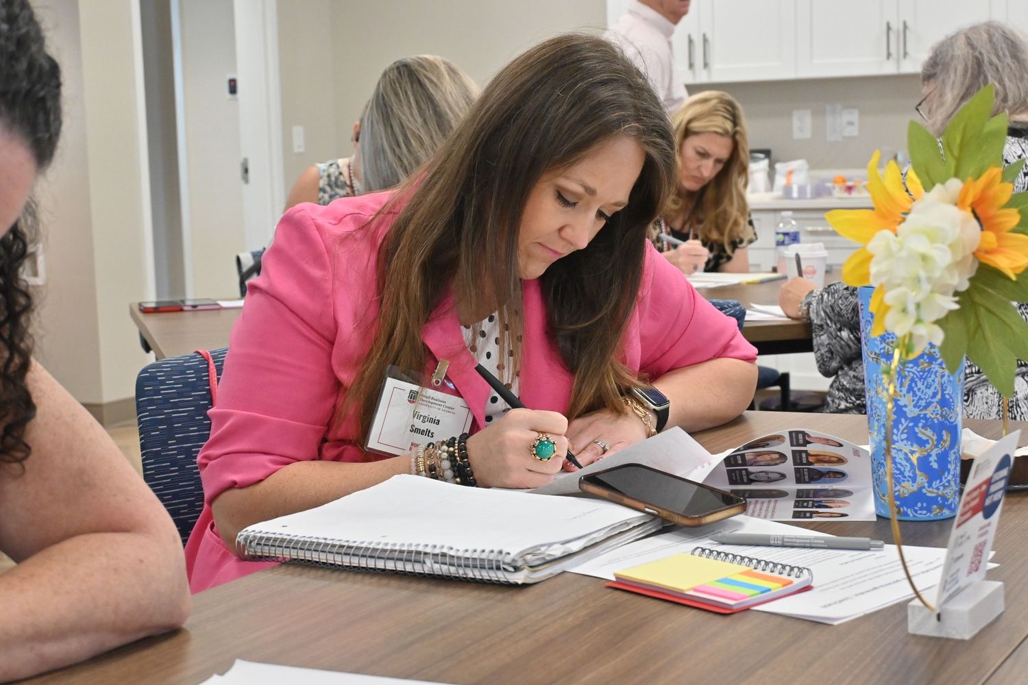 Woman in pink blazer writing in notebook at a table during a professional workshop or training session.