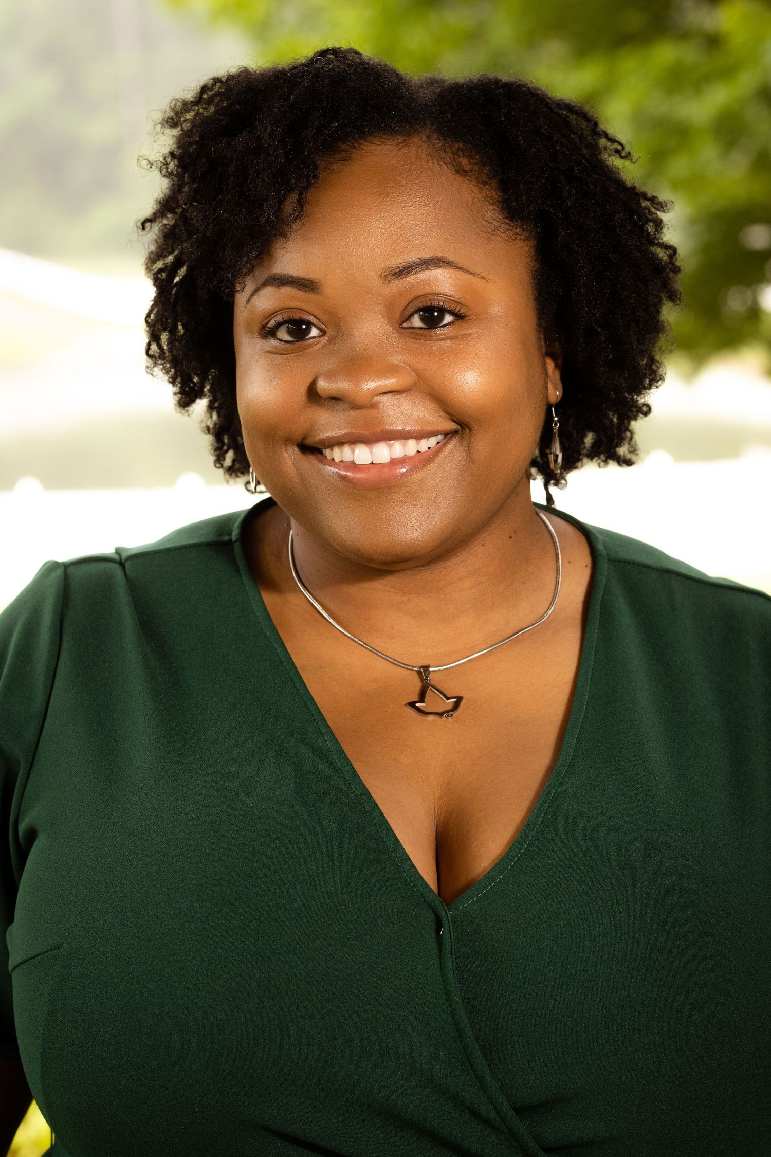 Smiling woman in green V-neck top with bird necklace, natural hair, outdoors with blurred green background.