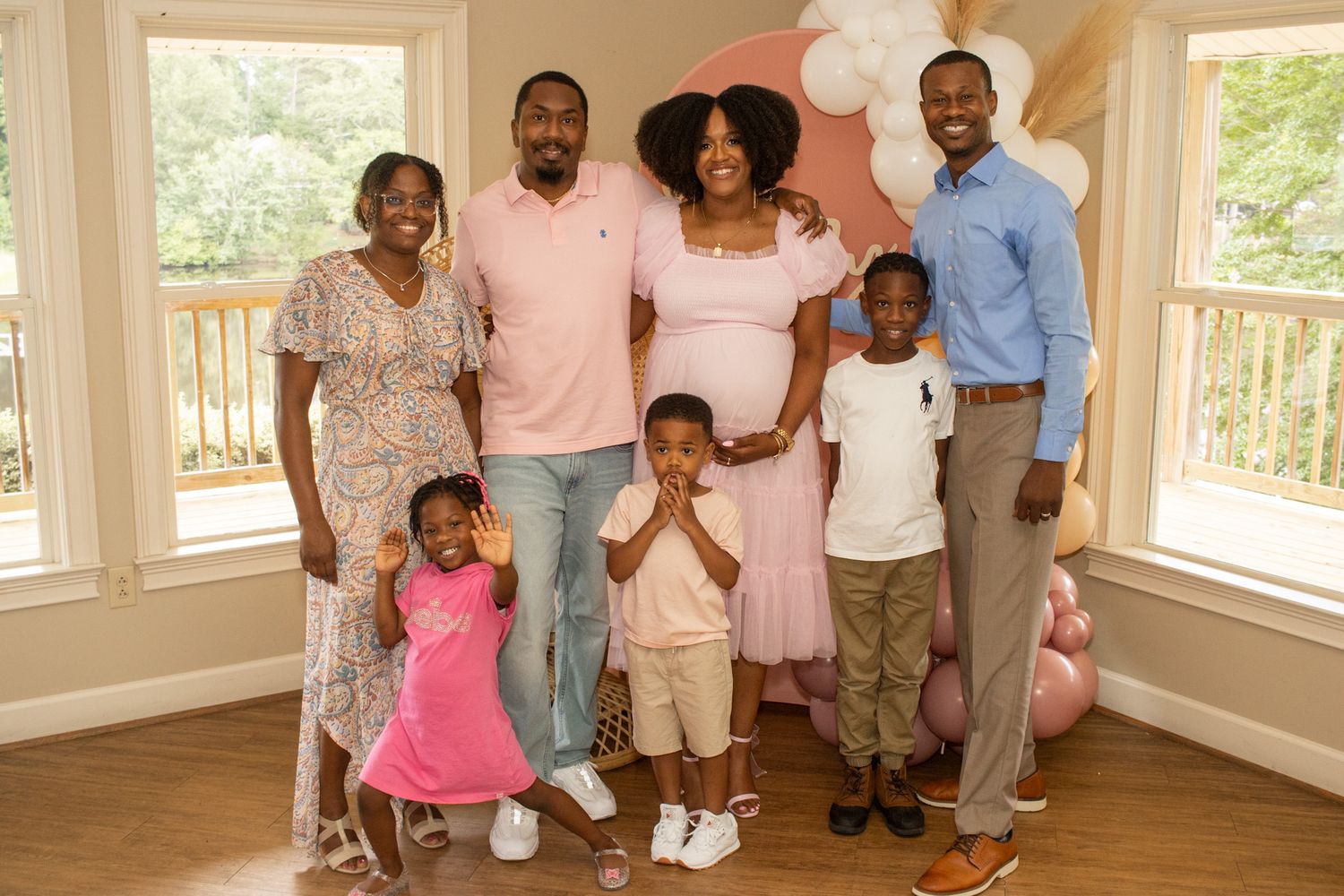 A family group poses together in a sunlit room with large windows and hardwood floors wearing coordinated pink and neutral outfits.