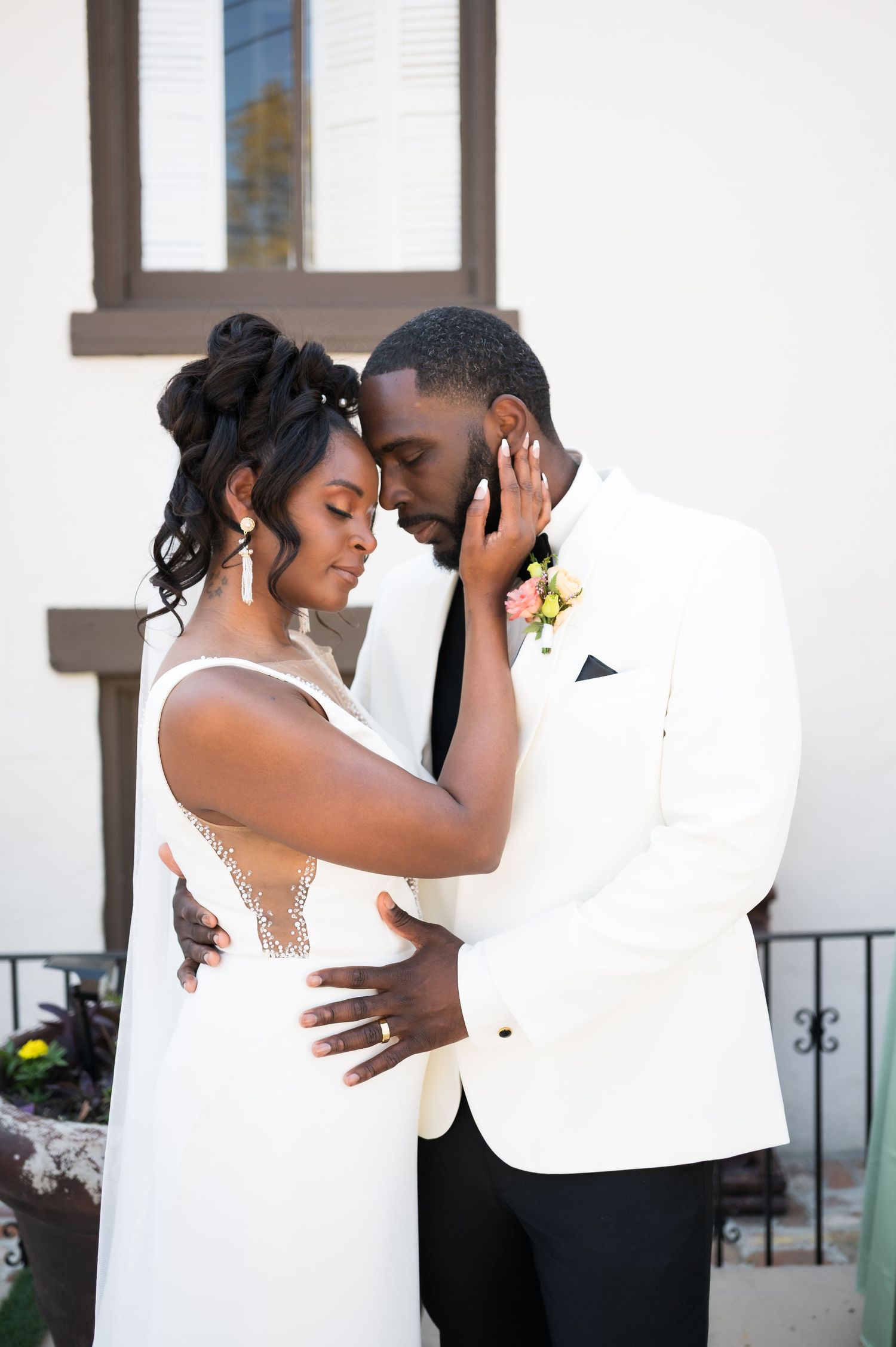 A Black bride and groom share an intimate forehead touch, dressed in elegant white wedding attire outdoors.