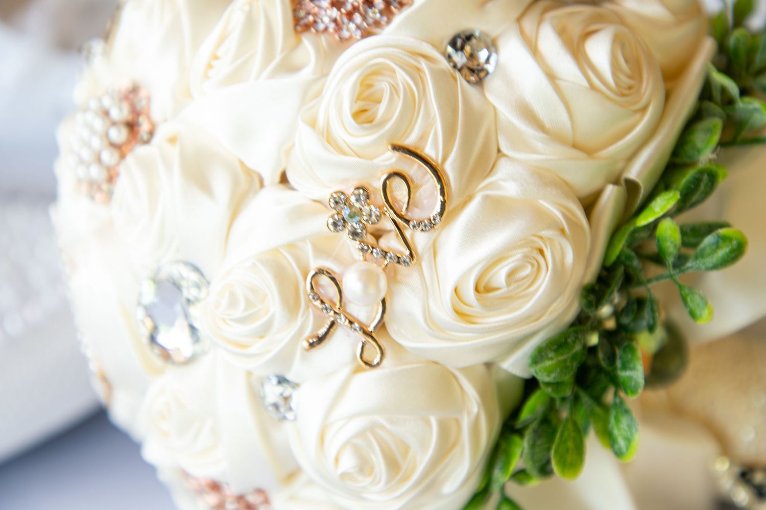 An elegant bridal bouquet of ivory satin roses adorned with gold script charms and sparkling brooch accents.