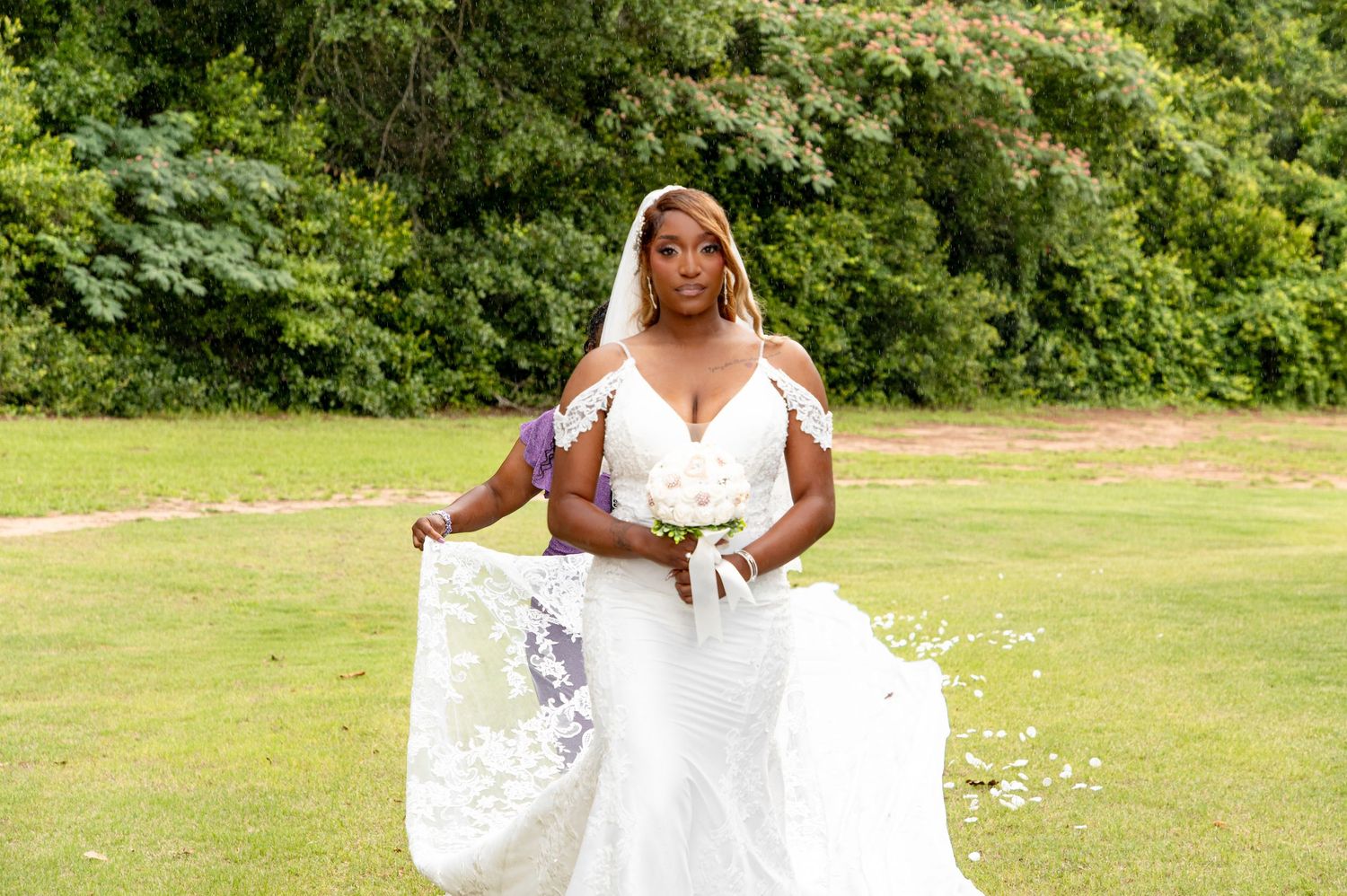 Bride in elegant white lace wedding gown holding bouquet and veil on green lawn.
