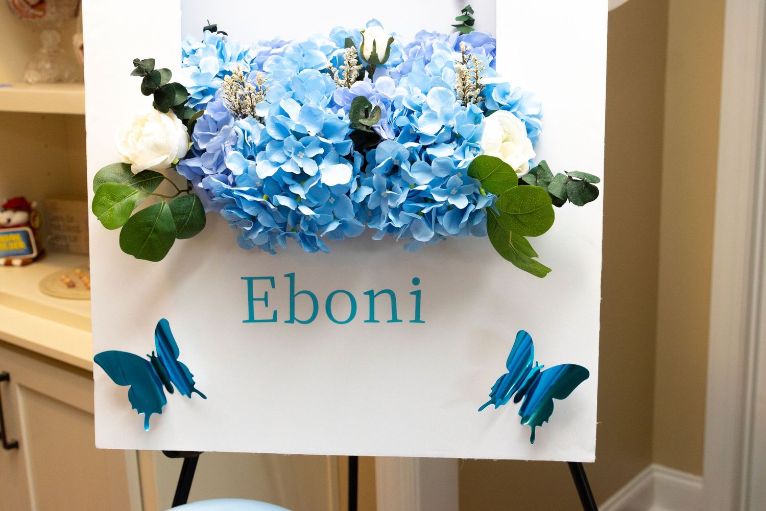 Handcrafted floral arrangement with vibrant blue hydrangeas and decorative butterfly accents displayed in white wall decor.