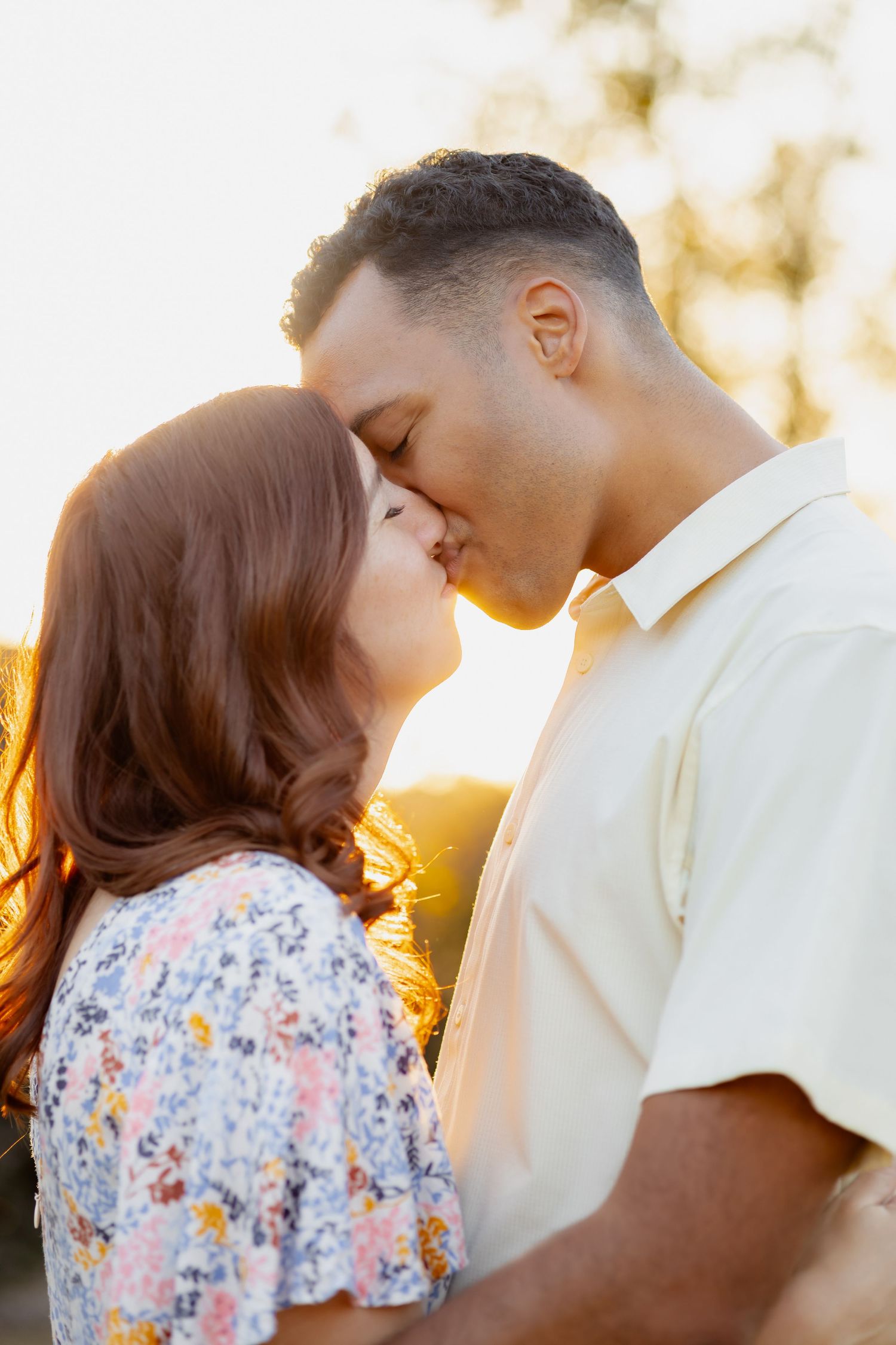 A couple shares an intimate romantic moment kissing during golden hour sunset in a dreamy outdoor setting.