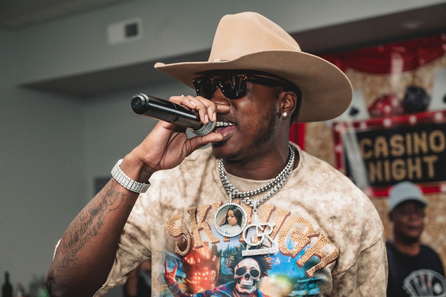 Person performs with microphone wearing a cowboy hat and patterned shirt at an indoor event.