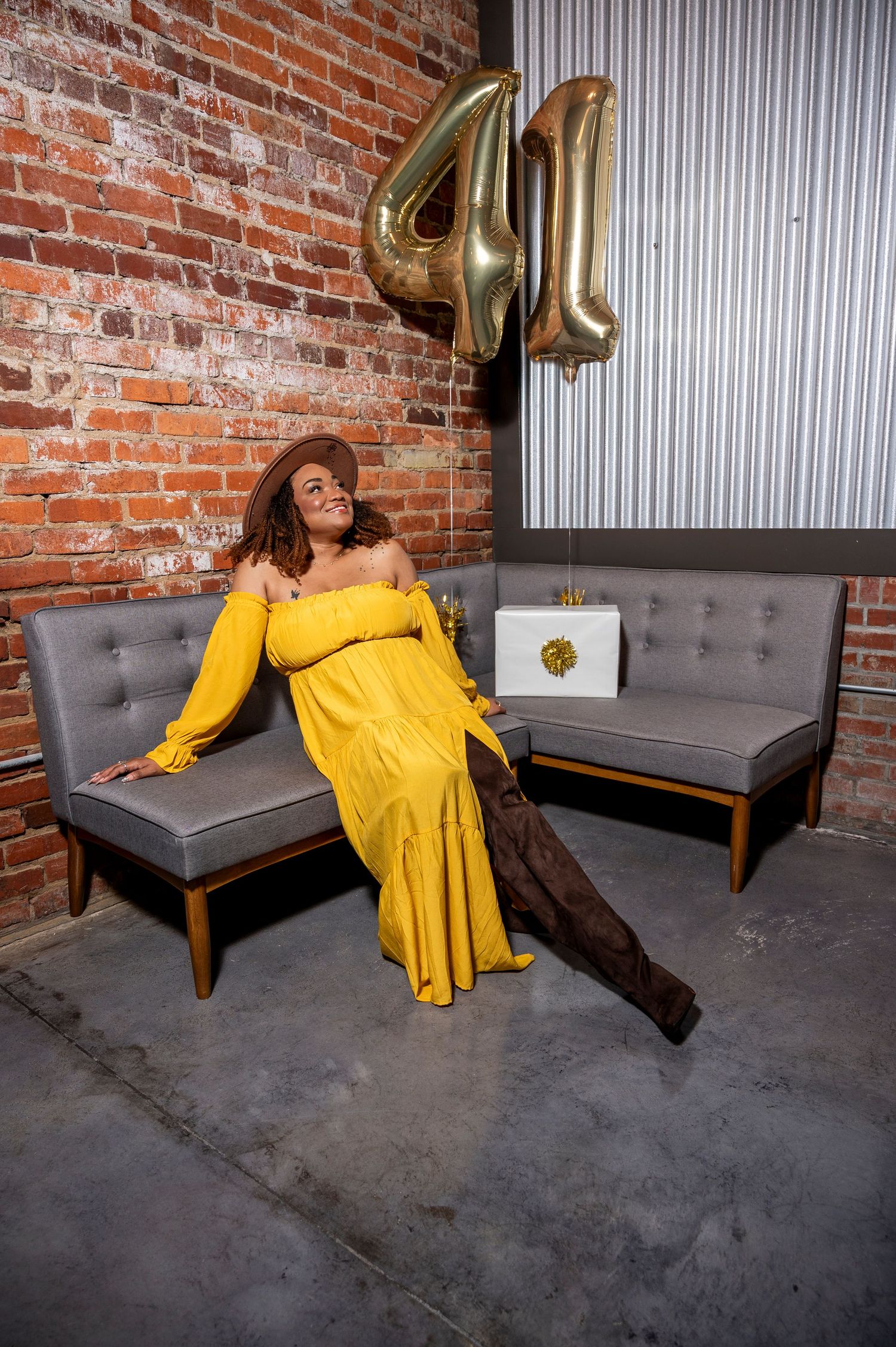 Woman in yellow off-shoulder gown poses on gray sofa with gold number 41 balloons against brick wall.