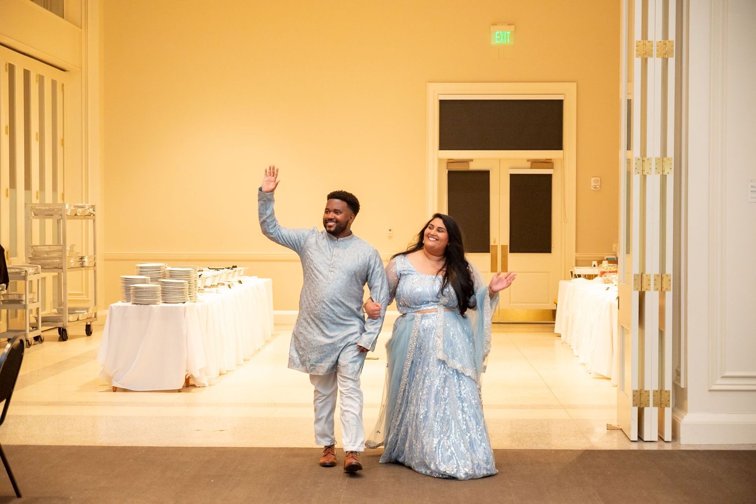 Couple in matching light blue Indian wedding attire waving as they enter an elegant banquet hall.