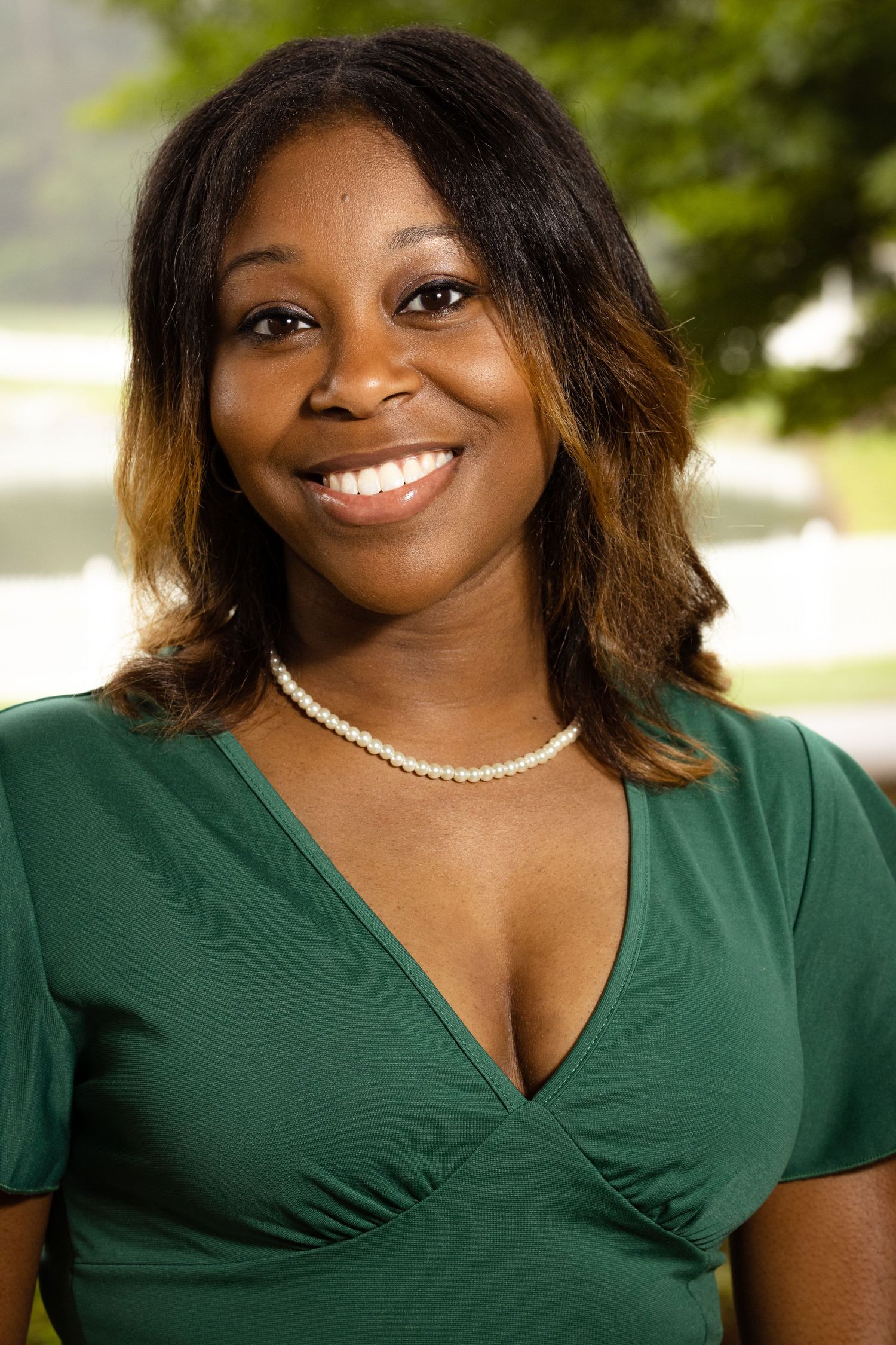 Smiling woman in green v-neck top and pearl necklace poses outdoors with lush greenery background.
