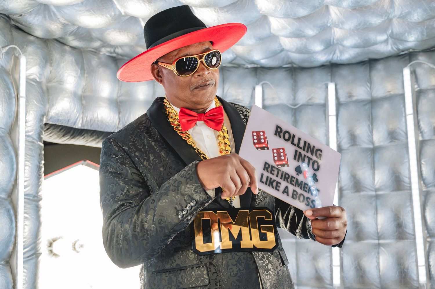 Concert performer in pink hat gestures while holding microphone against reflective silver backdrop.