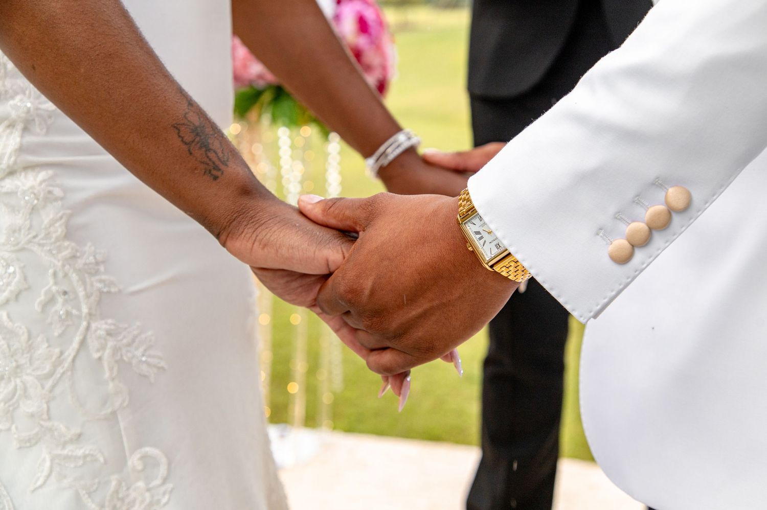 Bride and groom hold hands during wedding ceremony, groom wearing white suit with gold watch.