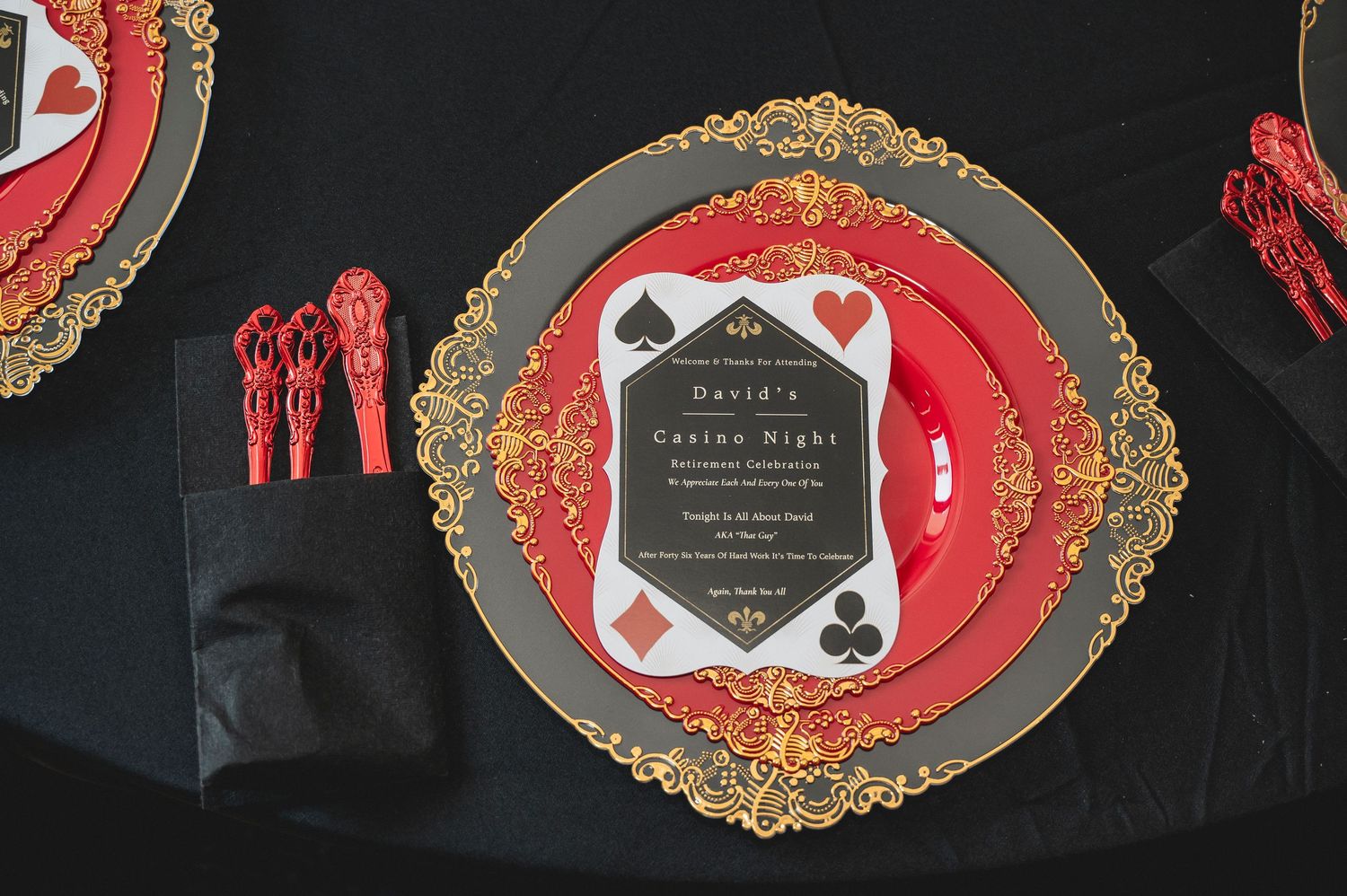 Elegant place setting with gold-rimmed charger plate, red plate, and black invitation card on black tablecloth.