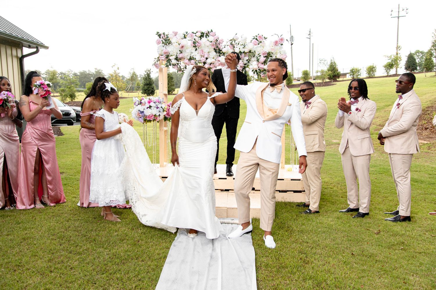 Joyful couple celebrates at outdoor wedding ceremony with bridal party in pink and tan.