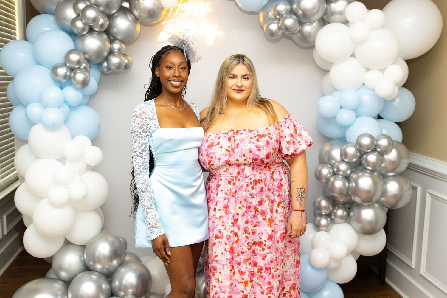 A festive scene with balloon decorations featuring a white and silver balloon arch and two people posing together.