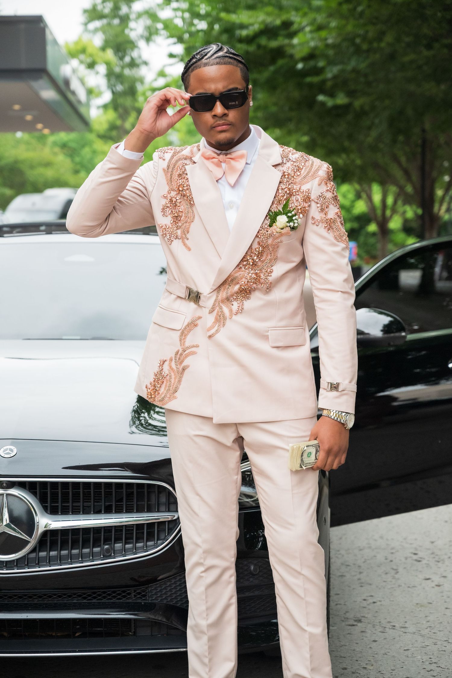 Young man in pink embroidered suit with bow tie and sunglasses posing in front of a Mercedes-Benz car.