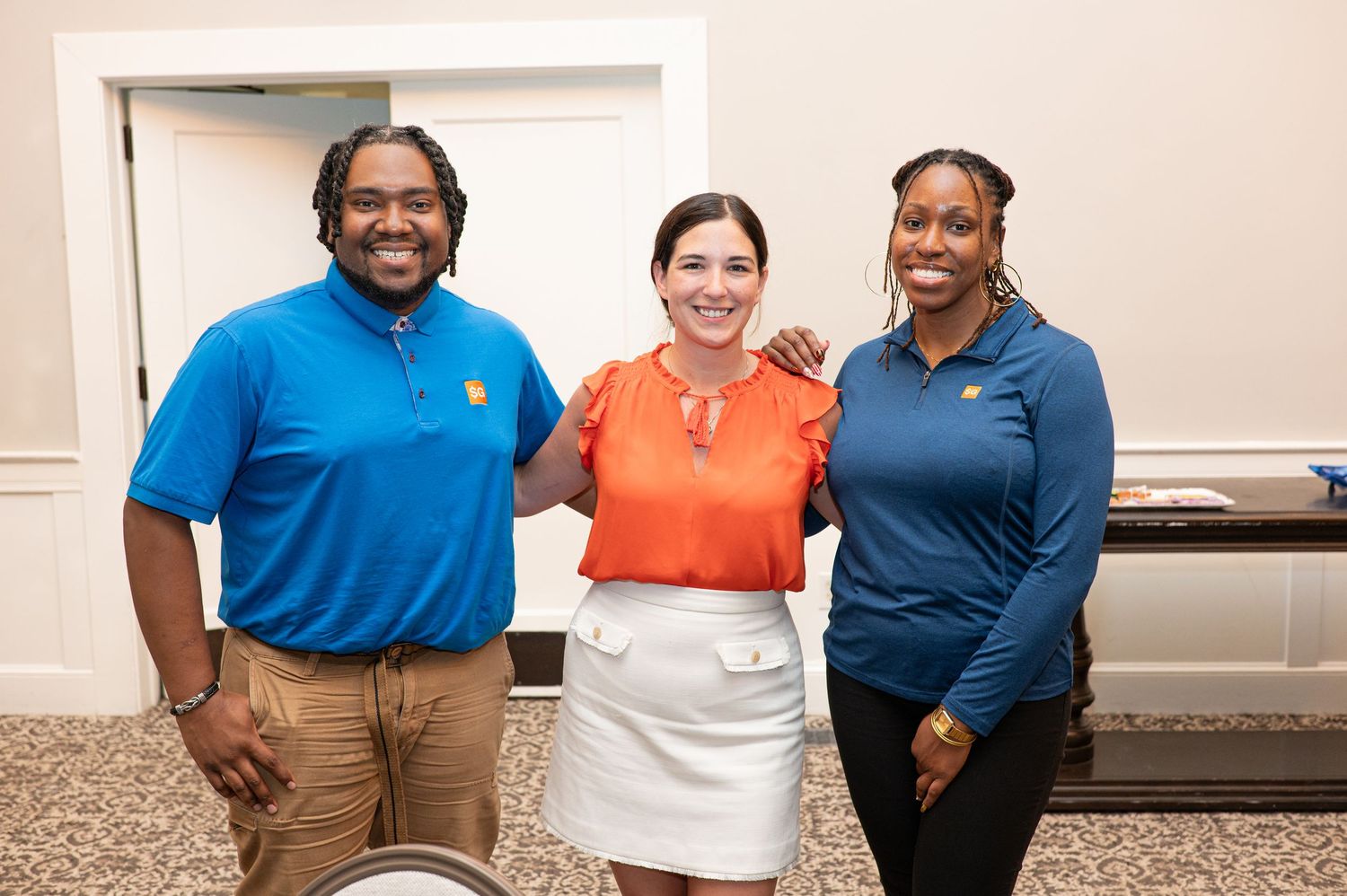 Three smiling colleagues pose together indoors, wearing blue and orange outfits at a professional event.