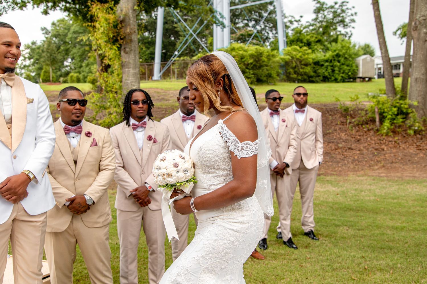 Bride walks past groomsmen in tan suits with burgundy ties at outdoor wedding.