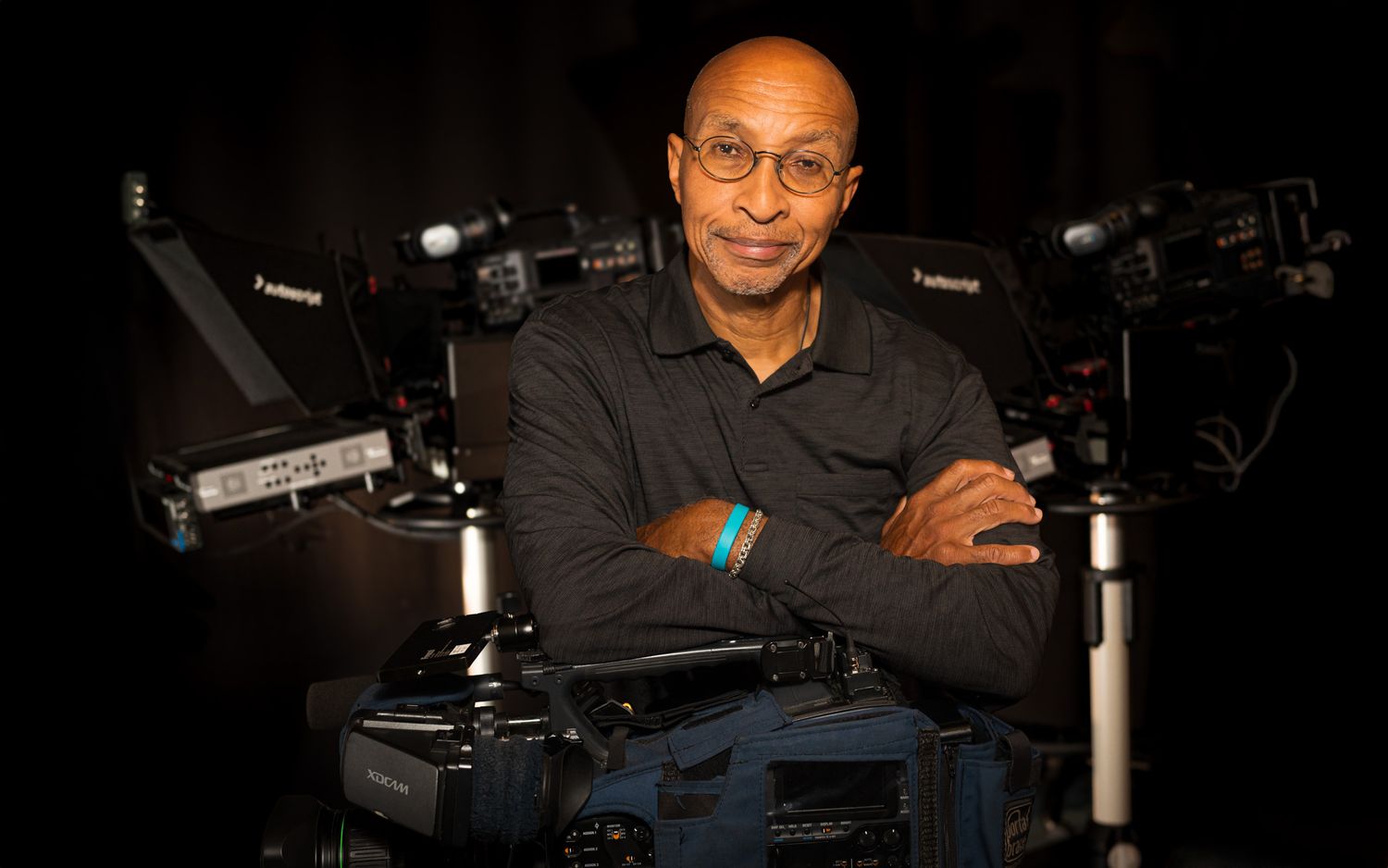 Man with glasses poses confidently with arms crossed beside professional video cameras in a dark studio setting.