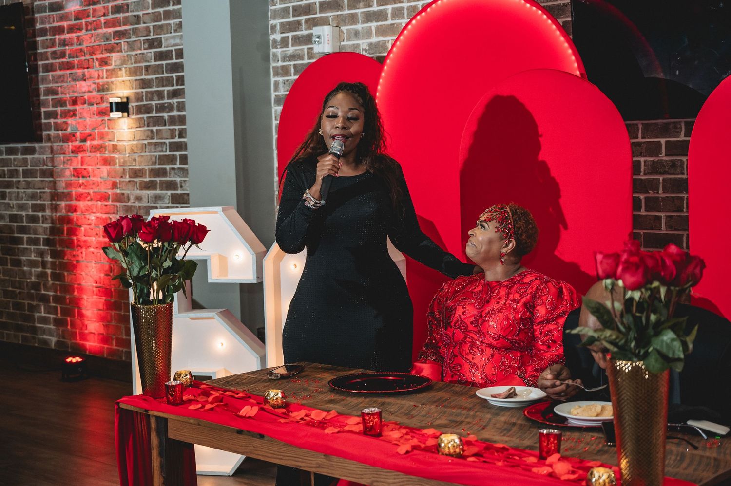 A romantic Valentine's Day themed party setup with red heart backdrop and roses on a decorated table.
