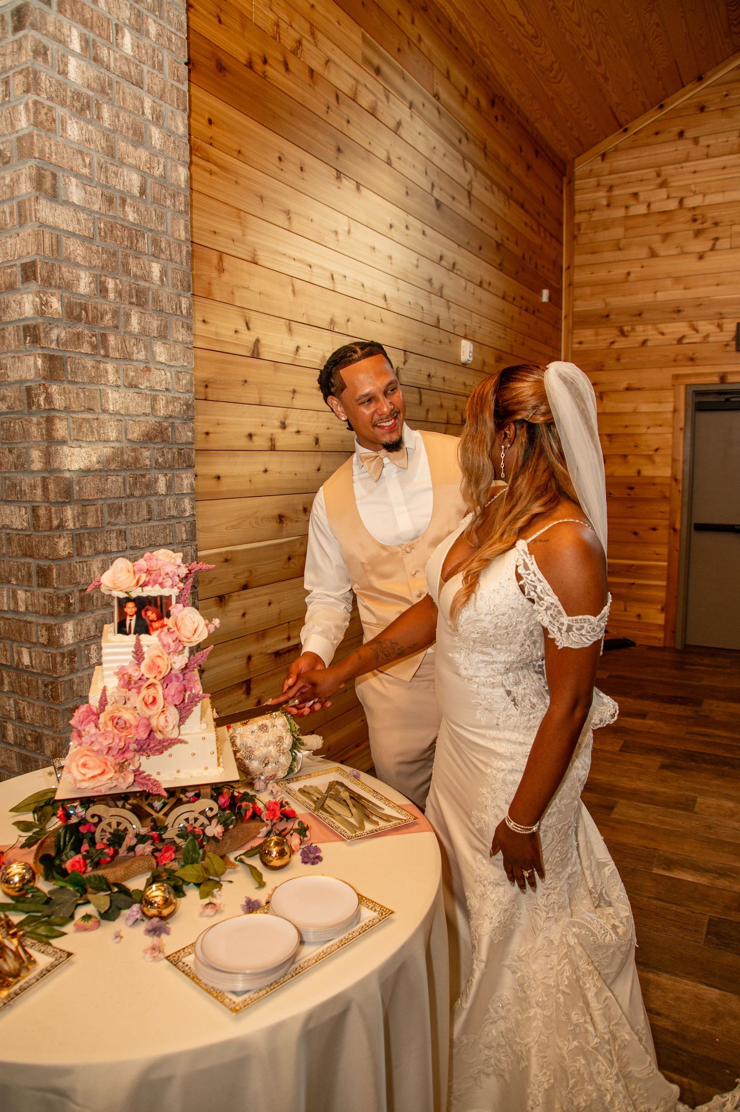 Bride and groom cutting tiered floral wedding cake in rustic wood-paneled venue.