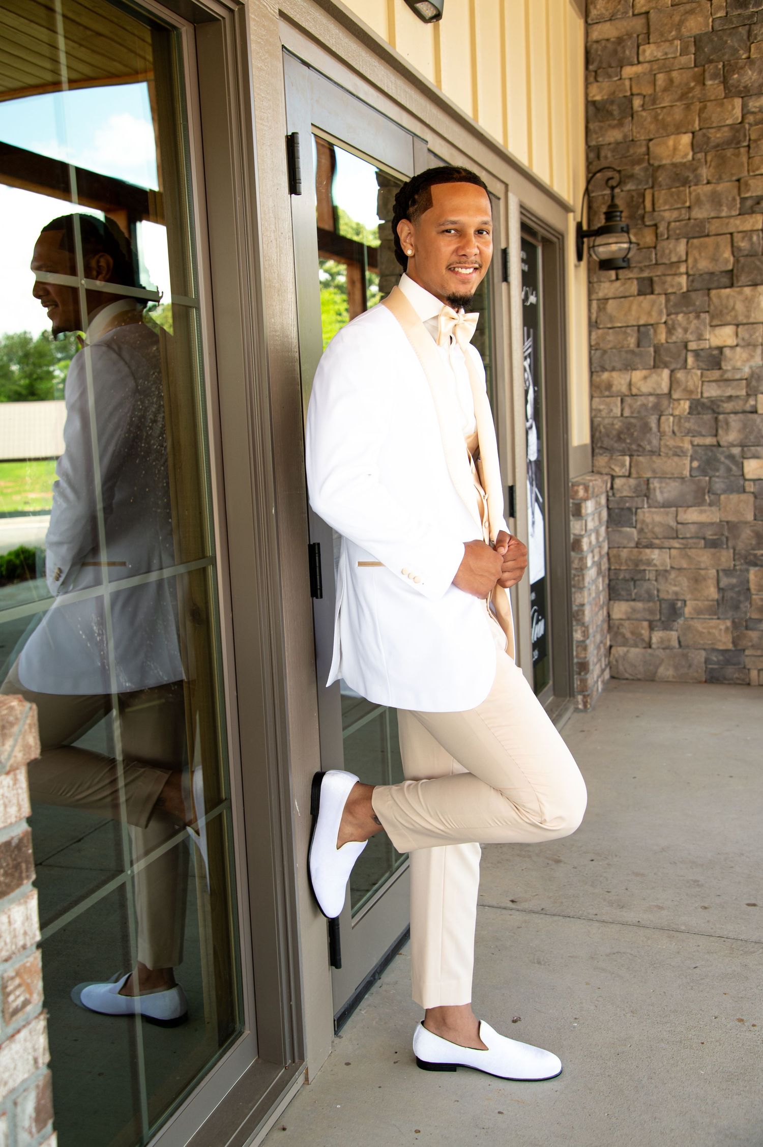 Groom in white blazer and khaki pants posing confidently against glass door at wedding venue.