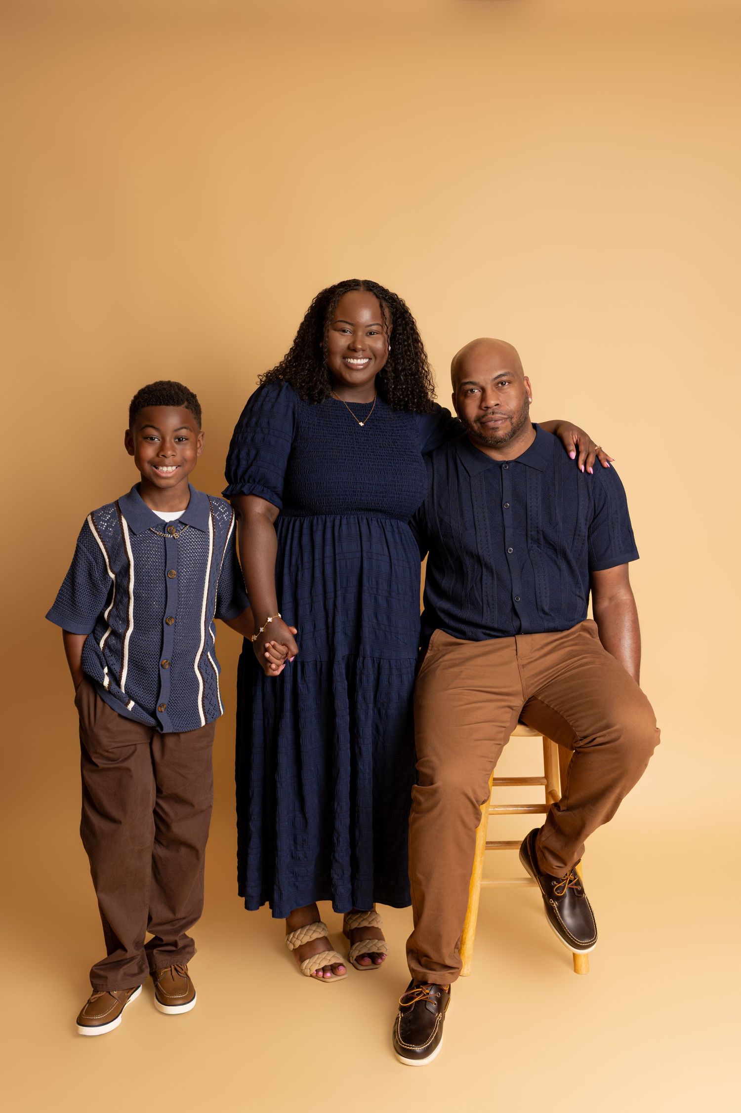 A family of three poses in a studio, wearing navy and brown outfits against a warm golden background.
