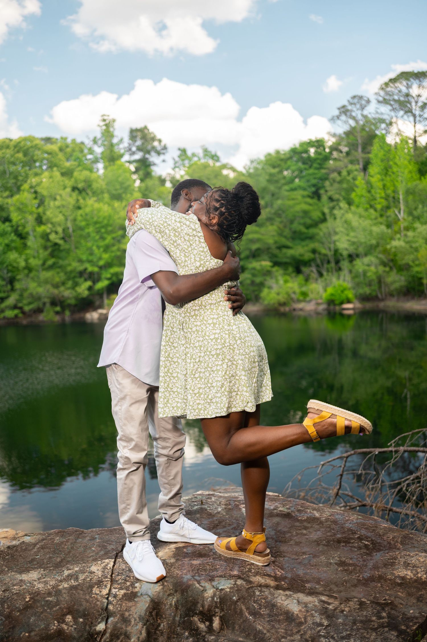 A couple embracing and kissing on rocks by a serene lake surrounded by lush green trees.