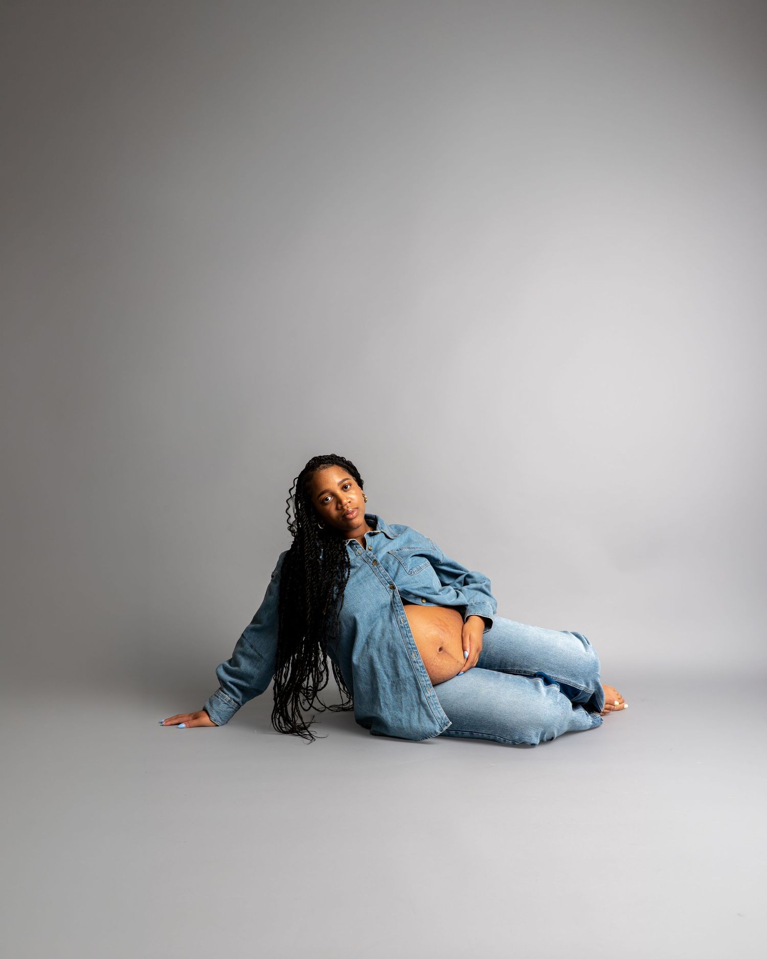 Pregnant woman in denim jacket and jeans sitting on studio floor showing baby bump with long braided hair.