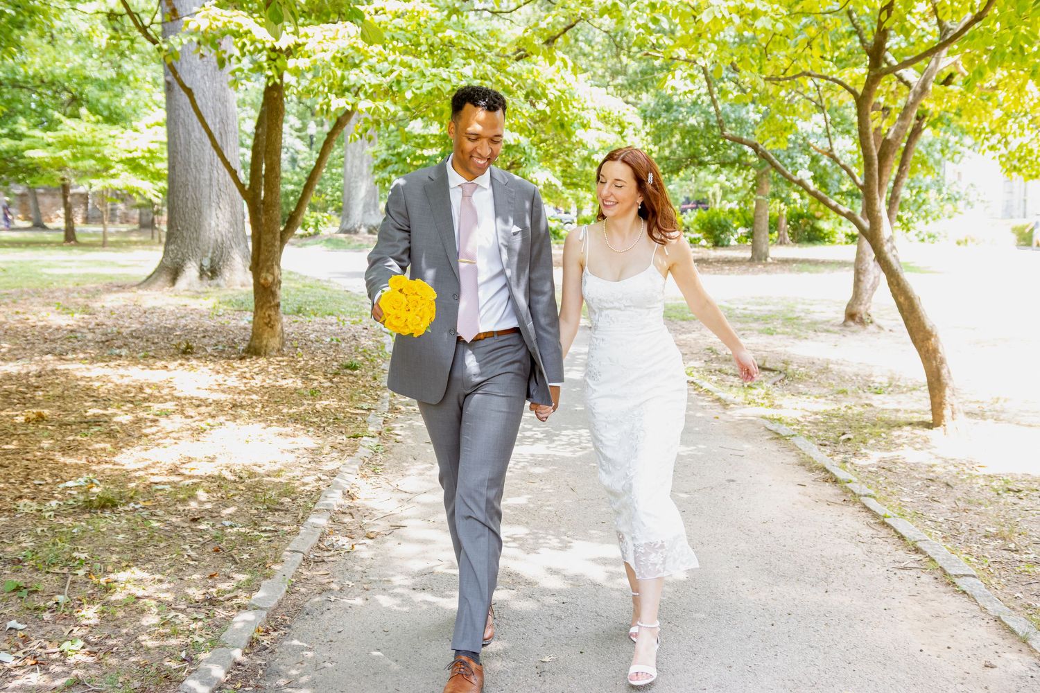 A couple in wedding attire walks hand in hand along a tree-lined path on a sunny day.