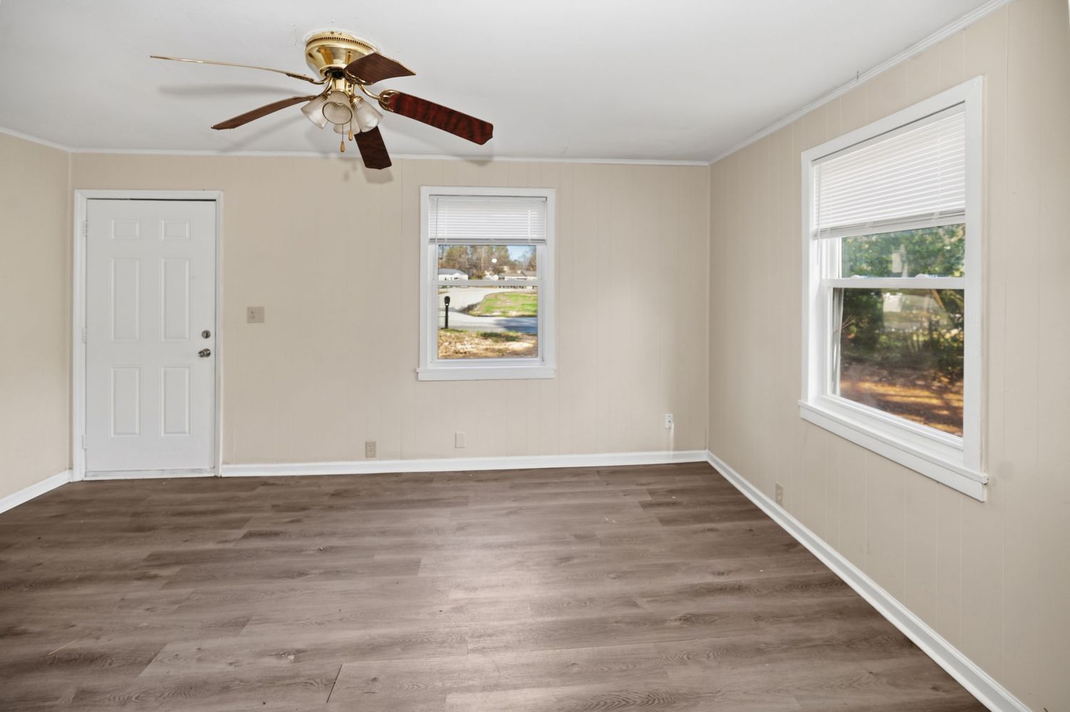 Bright empty living room with hardwood floors, ceiling fan, white trim, and two windows letting in natural light.