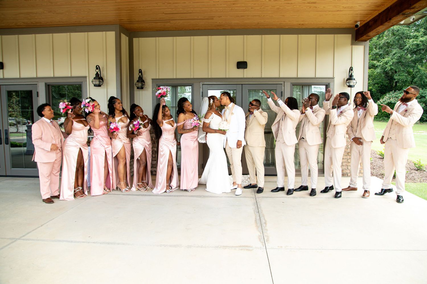 Large wedding party in pink bridesmaid dresses and white groomsmen suits pose under an outdoor pavilion.