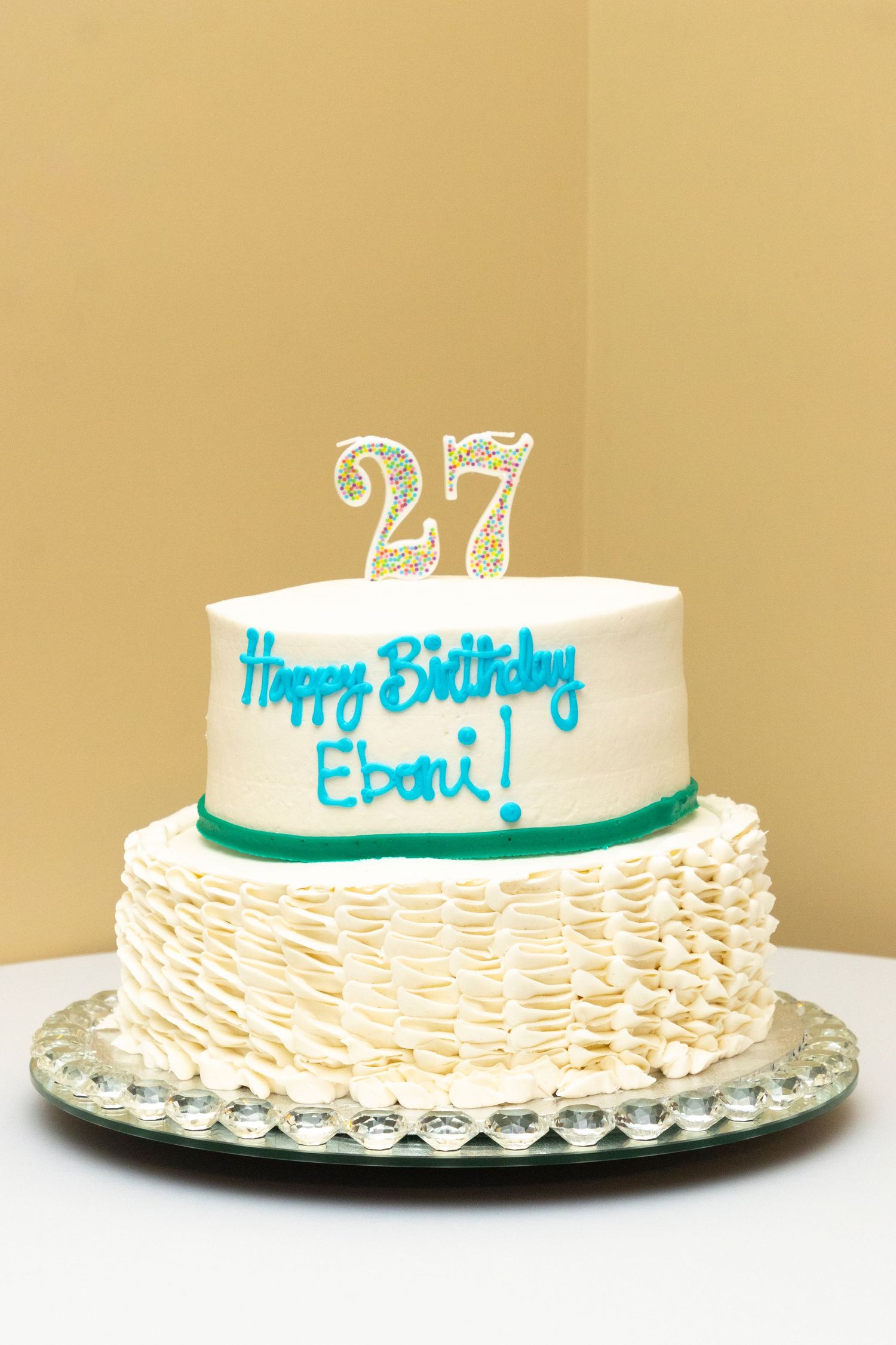 Birthday cake with white frosting and turquoise writing displayed from multiple angles on a glass cake plate.