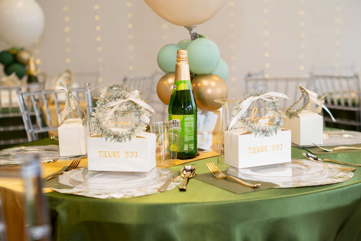 Elegant table setting with sage green tablecloth, white place cards, balloons and champagne bottle for celebration.