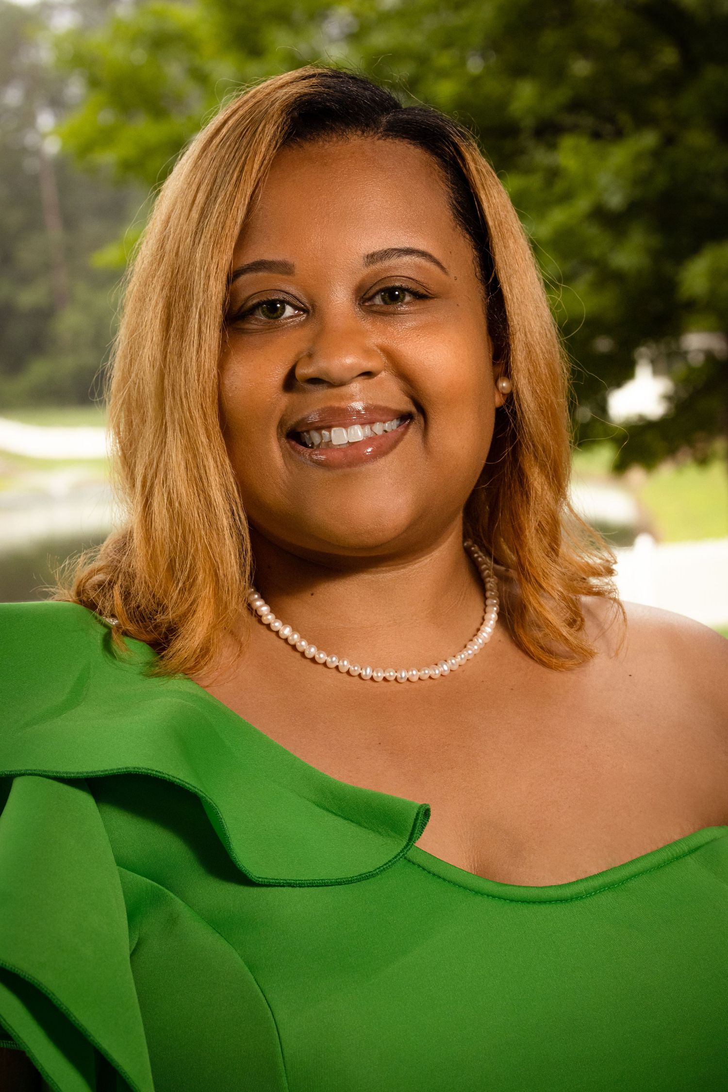Smiling woman in green ruffled top and pearl necklace with blonde highlights poses outdoors.